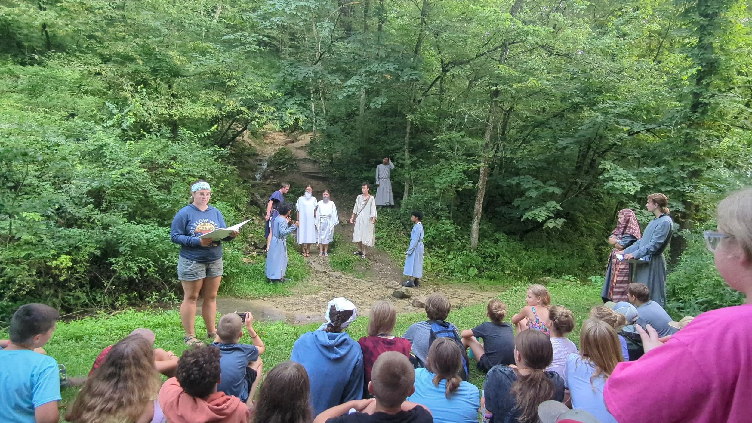 Group of people gathered outdoors in a lush green forest, some dressed in historical or biblical costumes, participating in an outdoor event or performance, with an audience of children and adults watching.