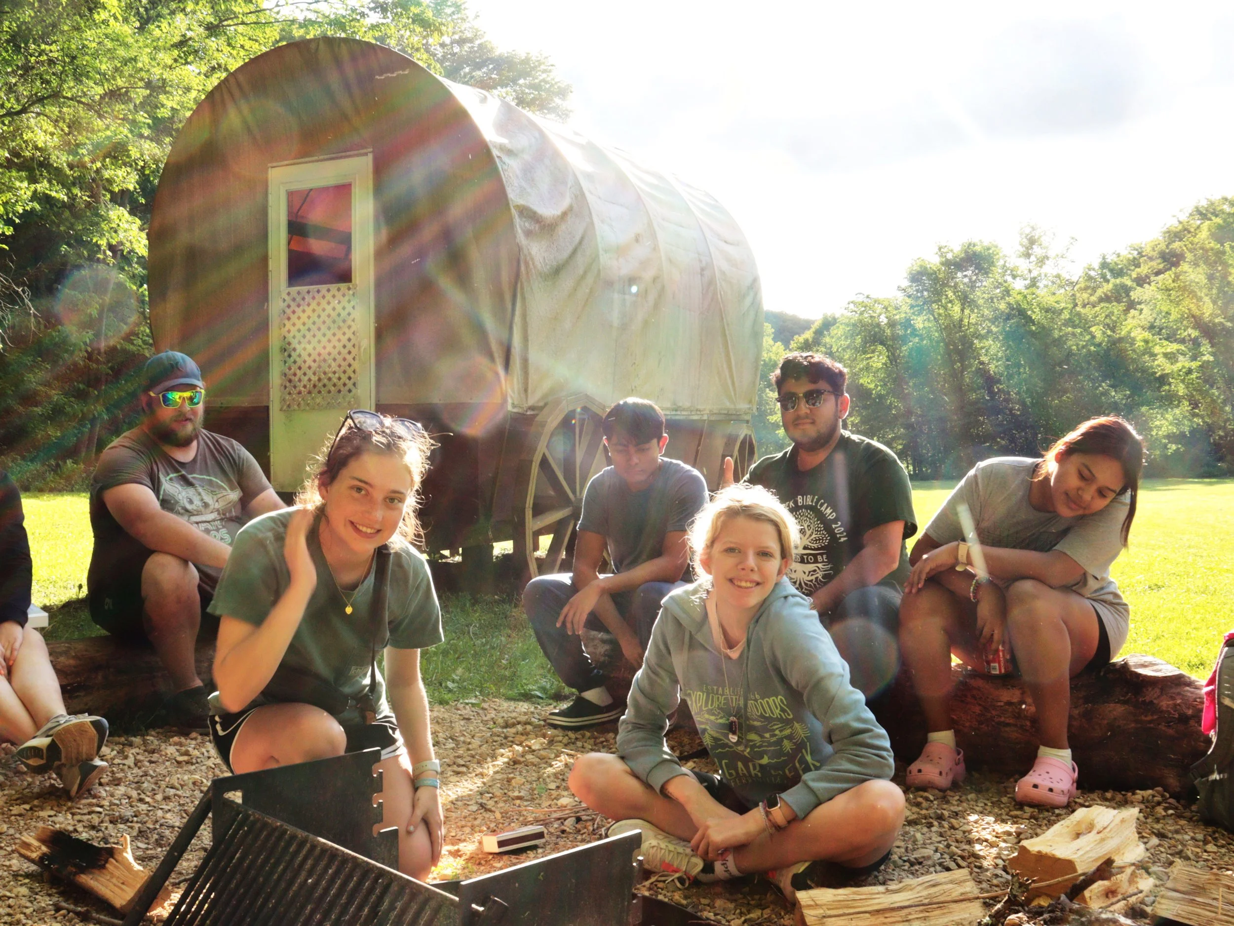 A group of people sitting outdoors in front of a covered wagon, surrounded by trees and sunlight.