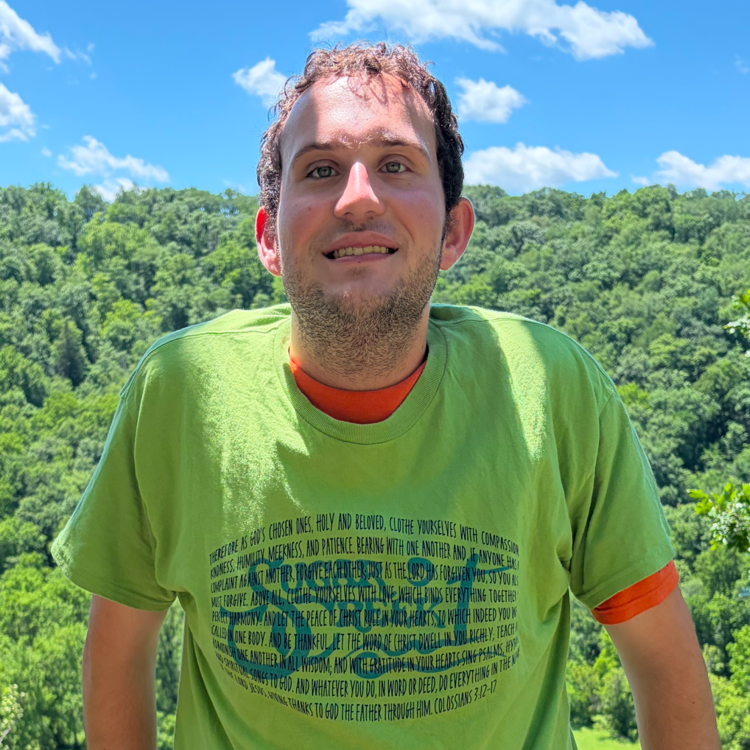 A young man wearing a bright green T-shirt with printed religious text, standing outdoors with a clear blue sky and lush green trees in the background.