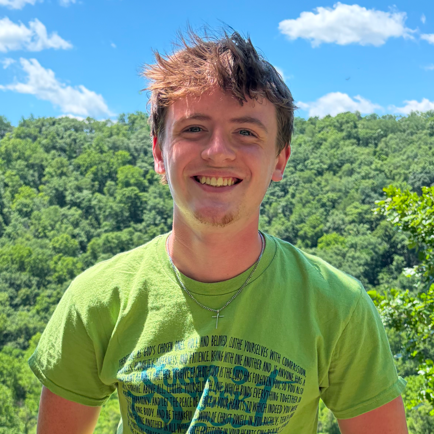 A young man with brown, messy hair, smiling and looking at the camera, standing outdoors with a background of green trees and a partly cloudy blue sky. He is wearing a bright green T-shirt with text and a silver cross necklace.