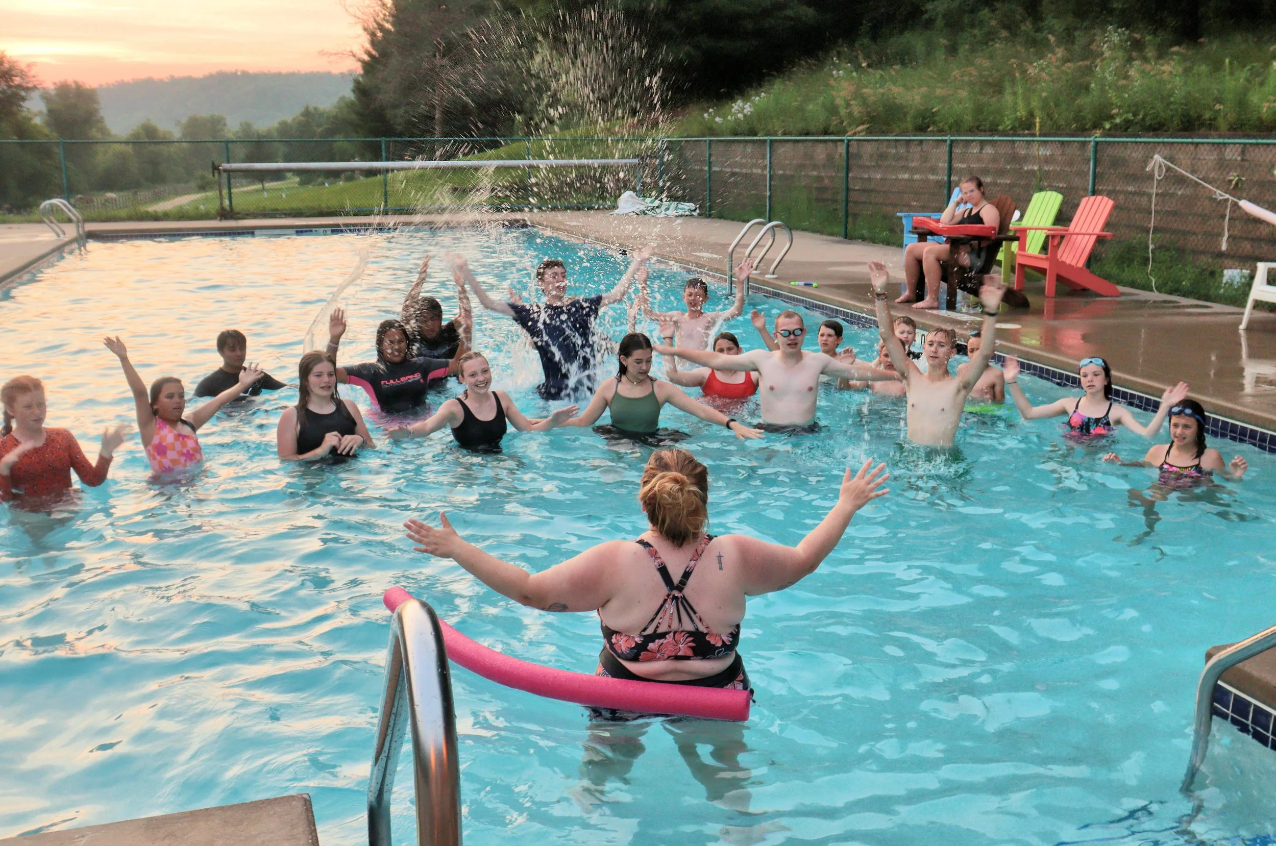 A group of children and teenagers are enjoying themselves in an outdoor swimming pool at sunset. Some are raising their hands in celebration, others are splashing water, and a woman with her back to the camera is sitting on the pool's edge, watching the scene.