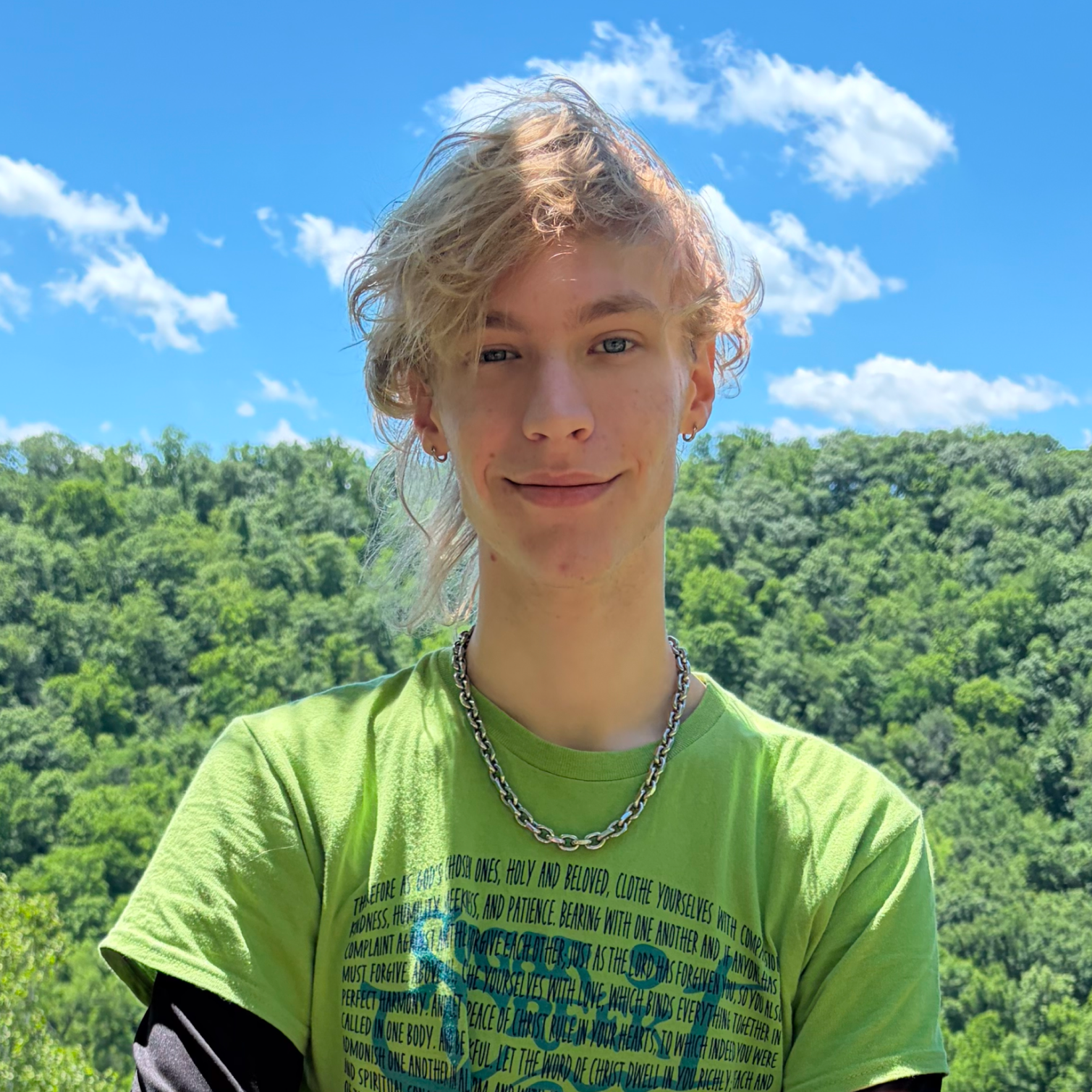 Young person with short, curly blonde hair, wearing a bright green t-shirt with scripture text on it, and a thick chain necklace, standing outdoors with a backdrop of lush green trees and a bright blue sky with white clouds.