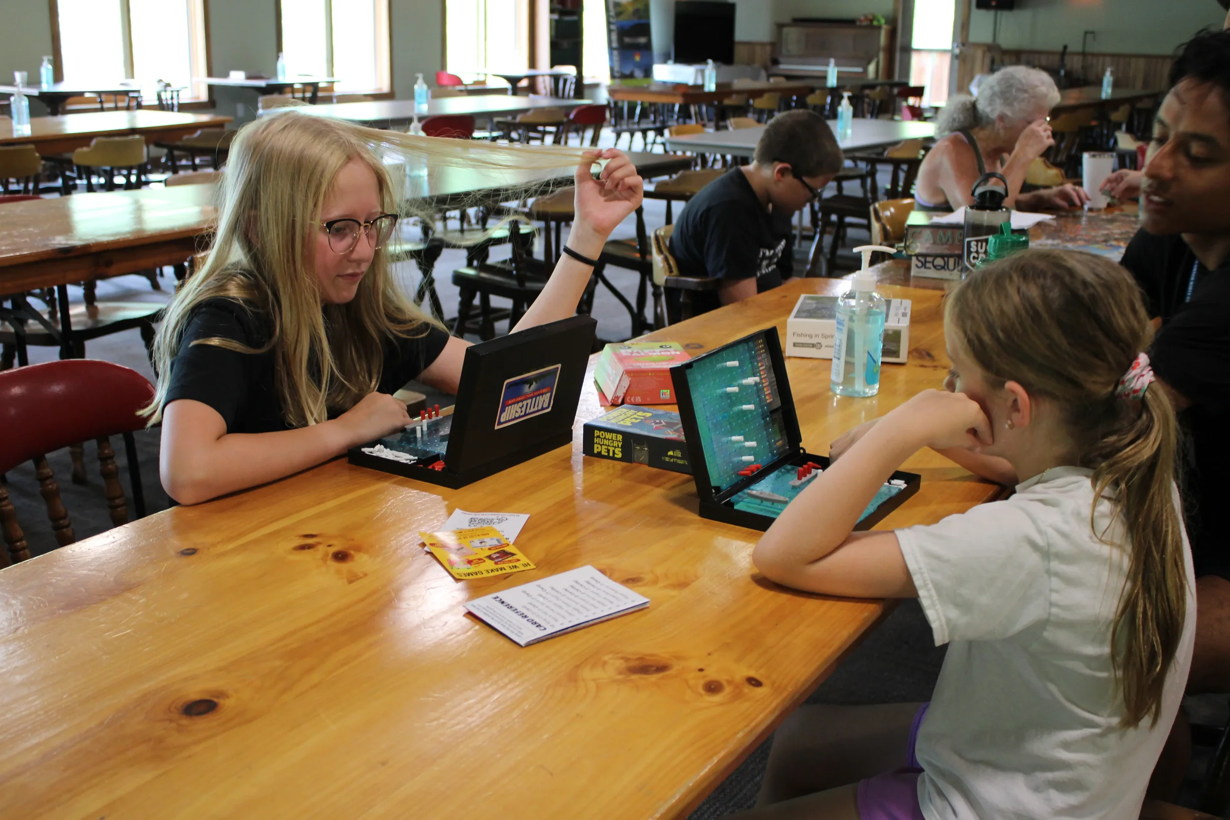 A group of children and adults playing board games and engaging in activities at a wooden table in a well-lit room with multiple empty tables and chairs, large windows, and wooden walls.