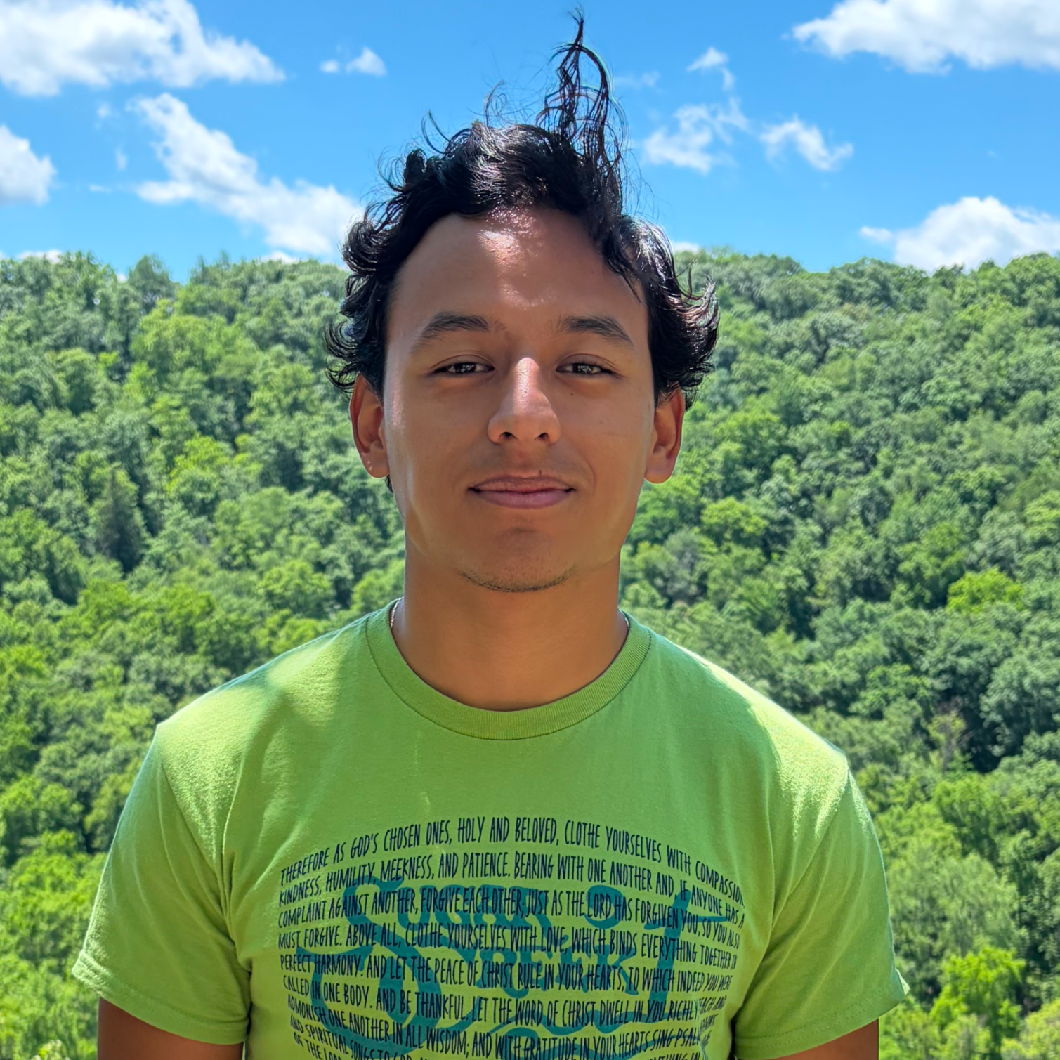 A young man with dark, wavy hair and a slight smile standing outdoors against a backdrop of lush green trees and a bright blue sky with white clouds.