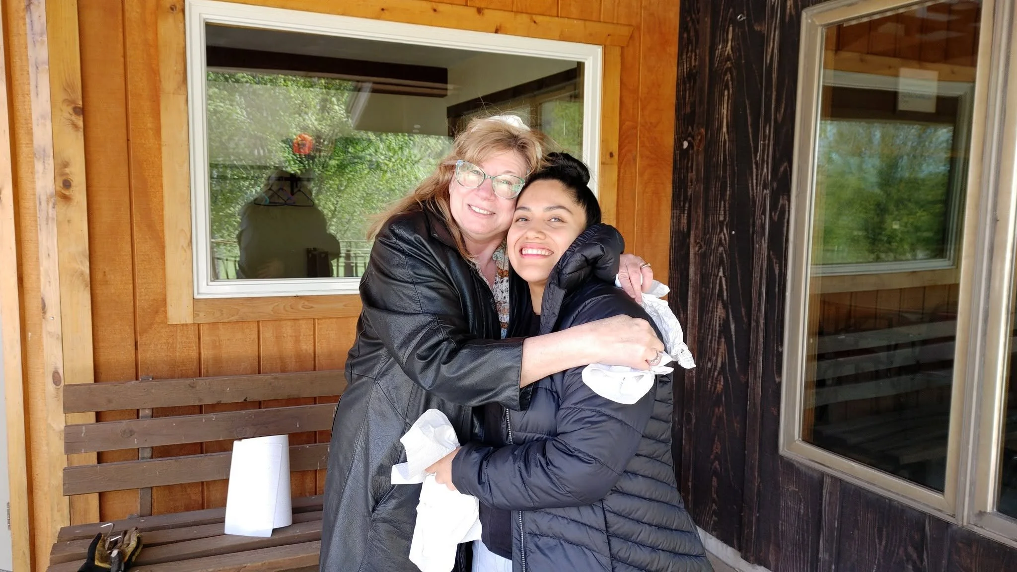 Two women hugging, smiling, standing outside in front of a house with wooden siding and large windows.