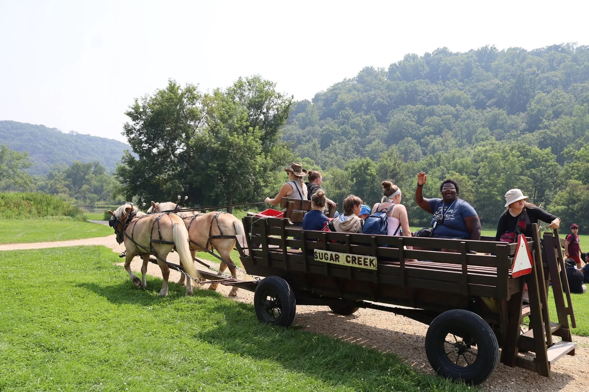 People riding horse-drawn wagon labeled 'SUGAR CREEK' through green park area with trees and hills in background, some waving and enjoying the ride.