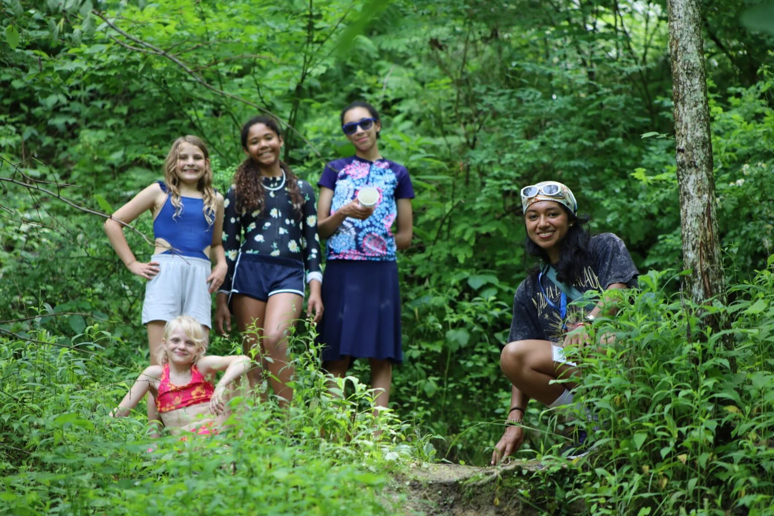 Group of five girls outdoors in a green forest, smiling and standing or crouching near a fallen tree