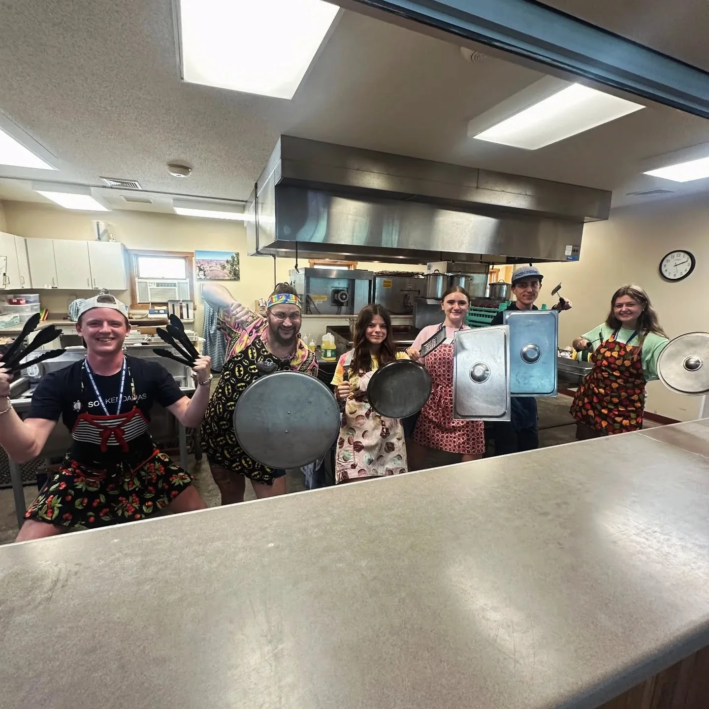 Group of six young people in a kitchen, wearing aprons and holding kitchen tools, smiling and posing for the camera.