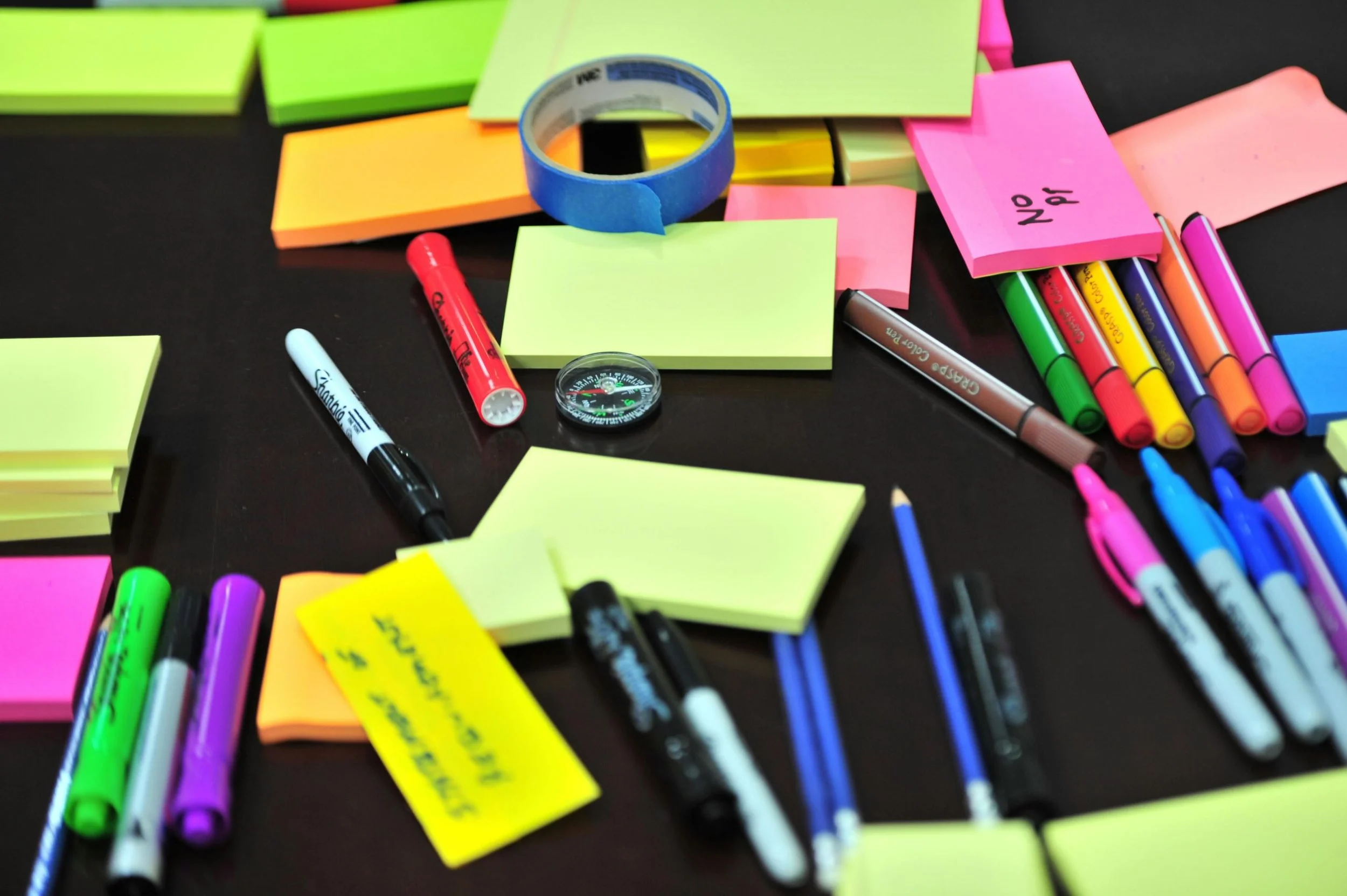 Colorful sticky notes, markers, tape, and a small compass scattered on a dark surface, indicating a planning or brainstorming session.