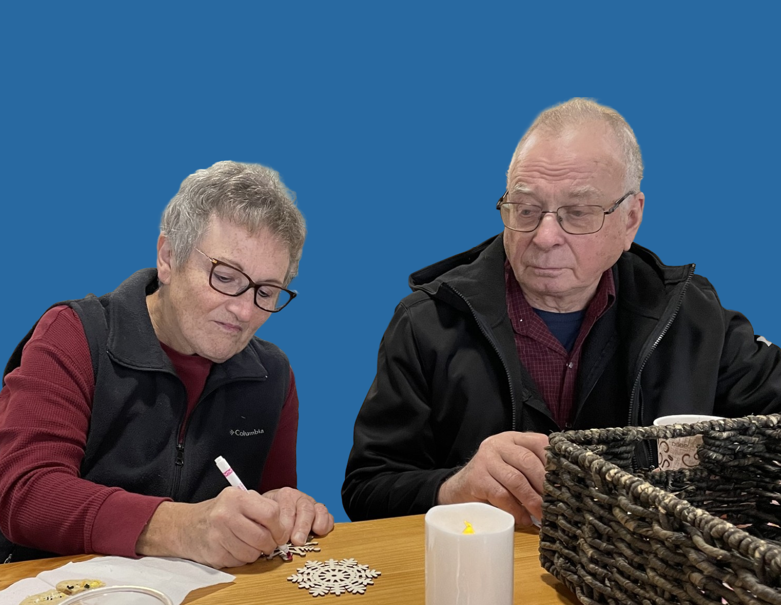 An elderly woman and man sitting at a table, decorating snowflake cutouts. The woman is writing or drawing, wearing glasses, a maroon shirt, and a black vest. The man, also wearing glasses, is holding a tray or basket, dressed in a black jacket over a red checkered shirt. There are candles and decorative items on the table.
