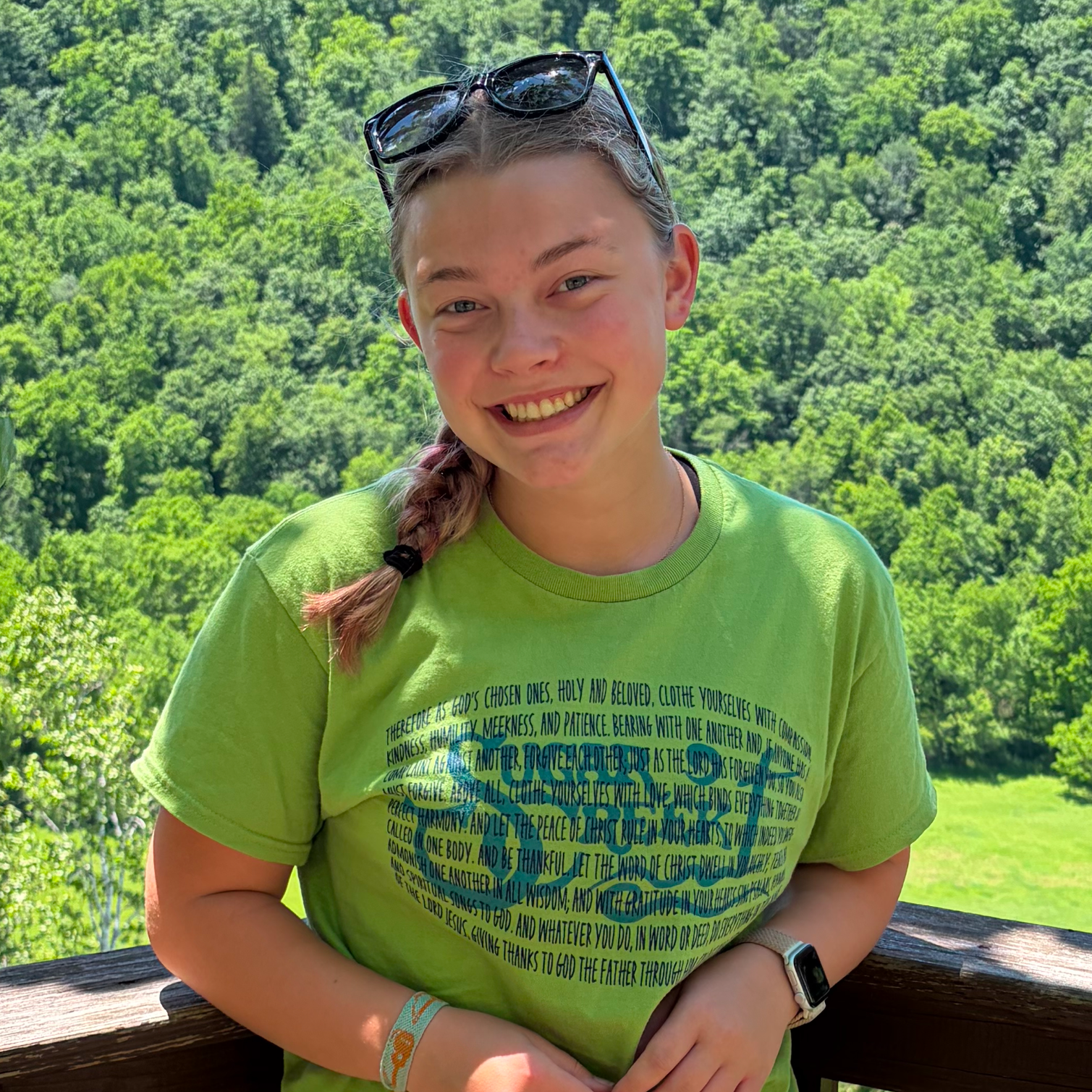 A young woman with glasses on her head, wearing a bright green T-shirt with a circular design and text, is smiling outdoors with a lush, green forest in the background. She has a braid over her shoulder and a smartwatch on her wrist.