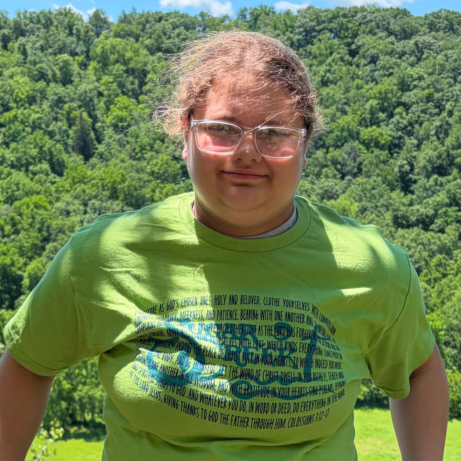 A person wearing glasses and a bright green T-shirt standing outdoors with a background of trees and a blue sky.