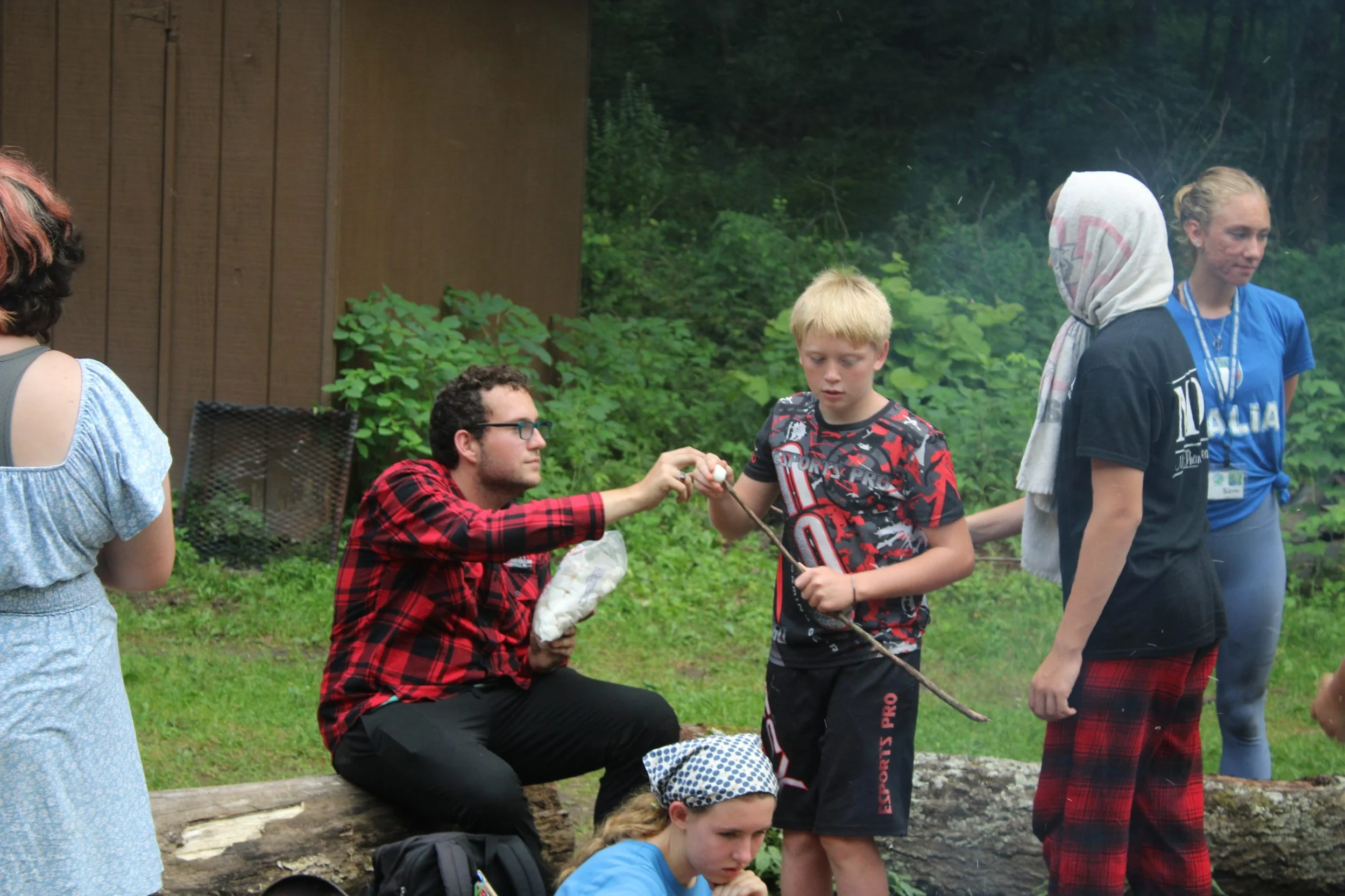 Group of children and teenagers outdoors, engaging in an activity involving a stick and a bag, in a wooded area with green foliage.