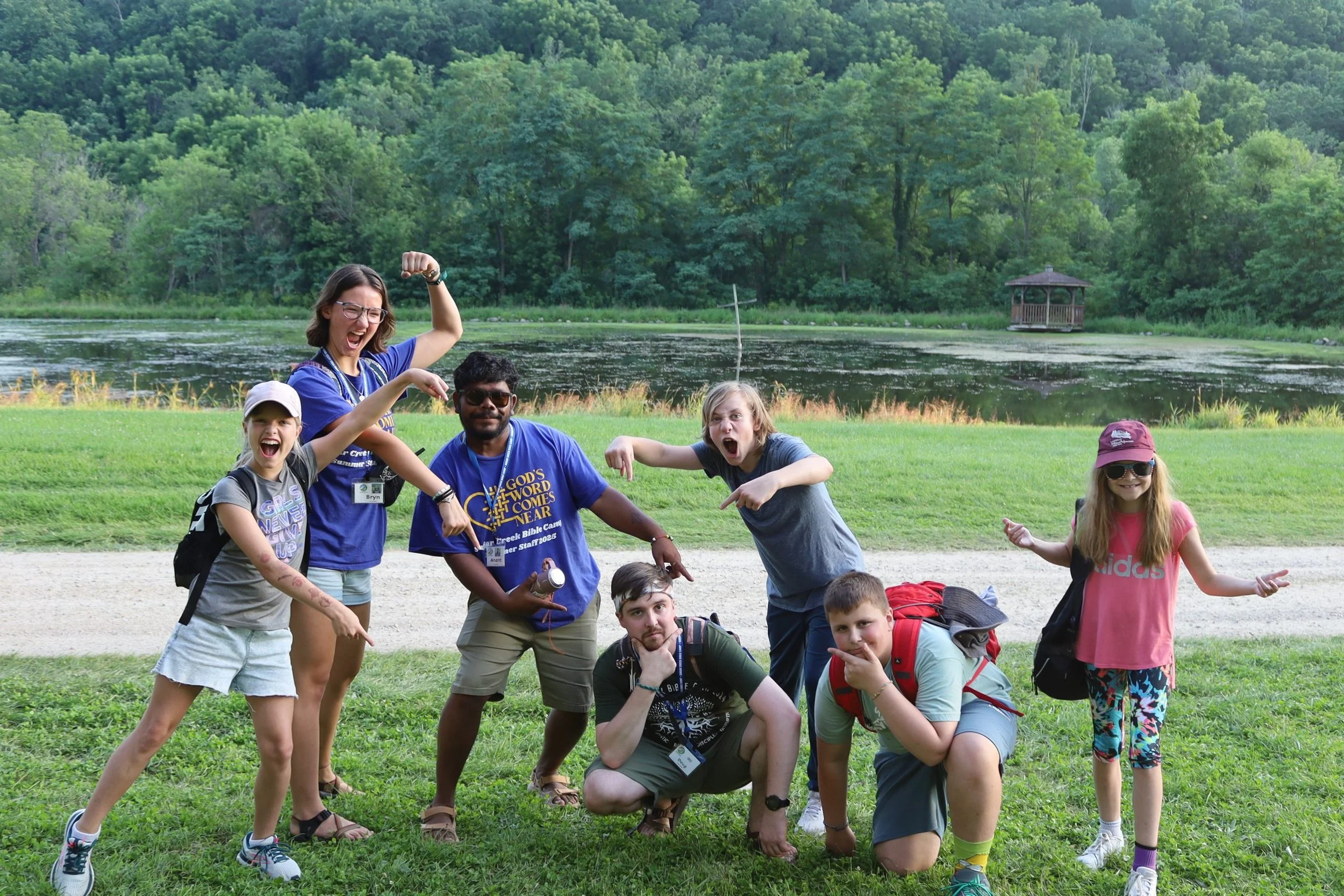 Group of children and a woman outdoors near a lake, posing playfully for a photo with lush trees and a gazebo in the background.