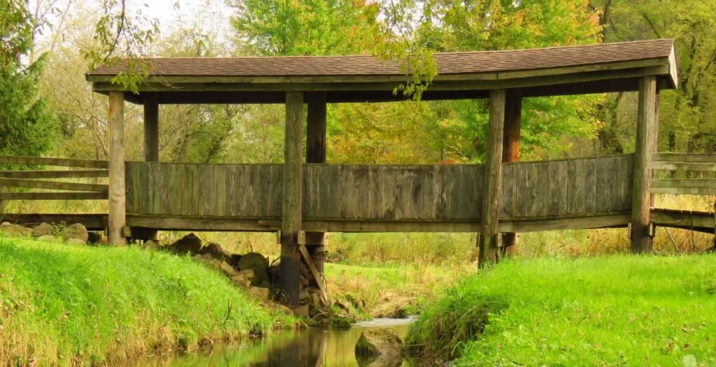 A wooden covered bridge over a small stream in a green park with trees.