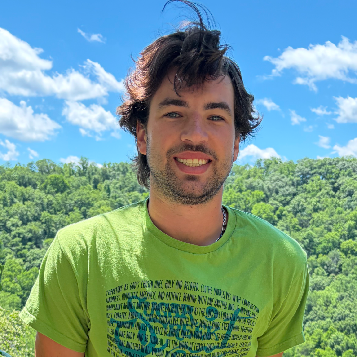 A young man with dark, wavy hair and light skin smiling outdoors against a green, wooded landscape and blue sky with clouds. He is wearing a bright green T-shirt with text and a silver chain necklace.
