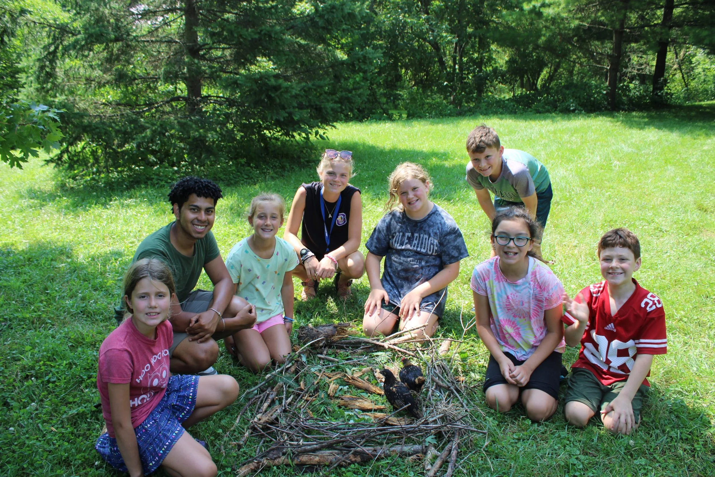 Group of children and a young adult sitting and kneeling on grass in a wooded area, posing with a small campfire made of sticks and bird nests, smiling at the camera.