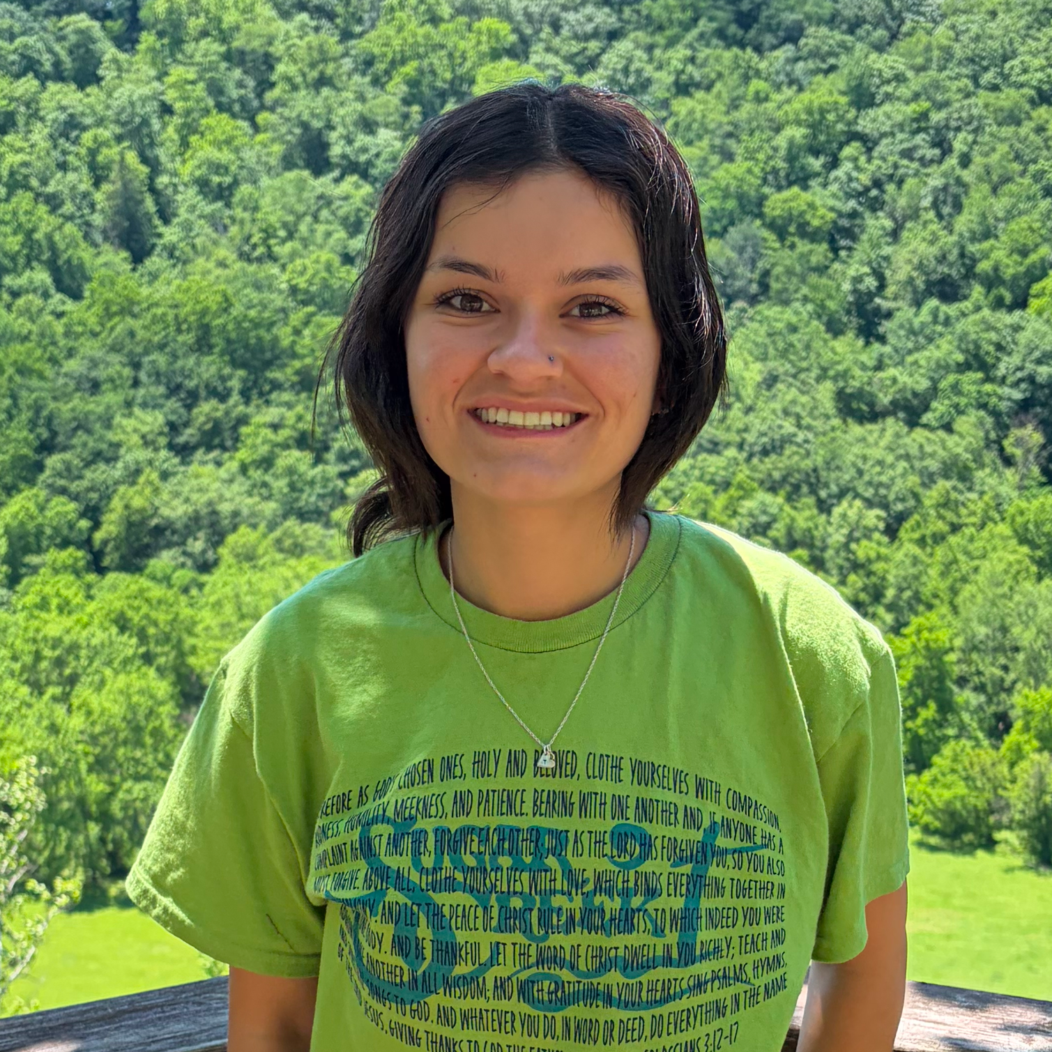 A young woman with short dark hair, wearing a green T-shirt and necklace, smiling outdoors with a green, forested hillside in the background.