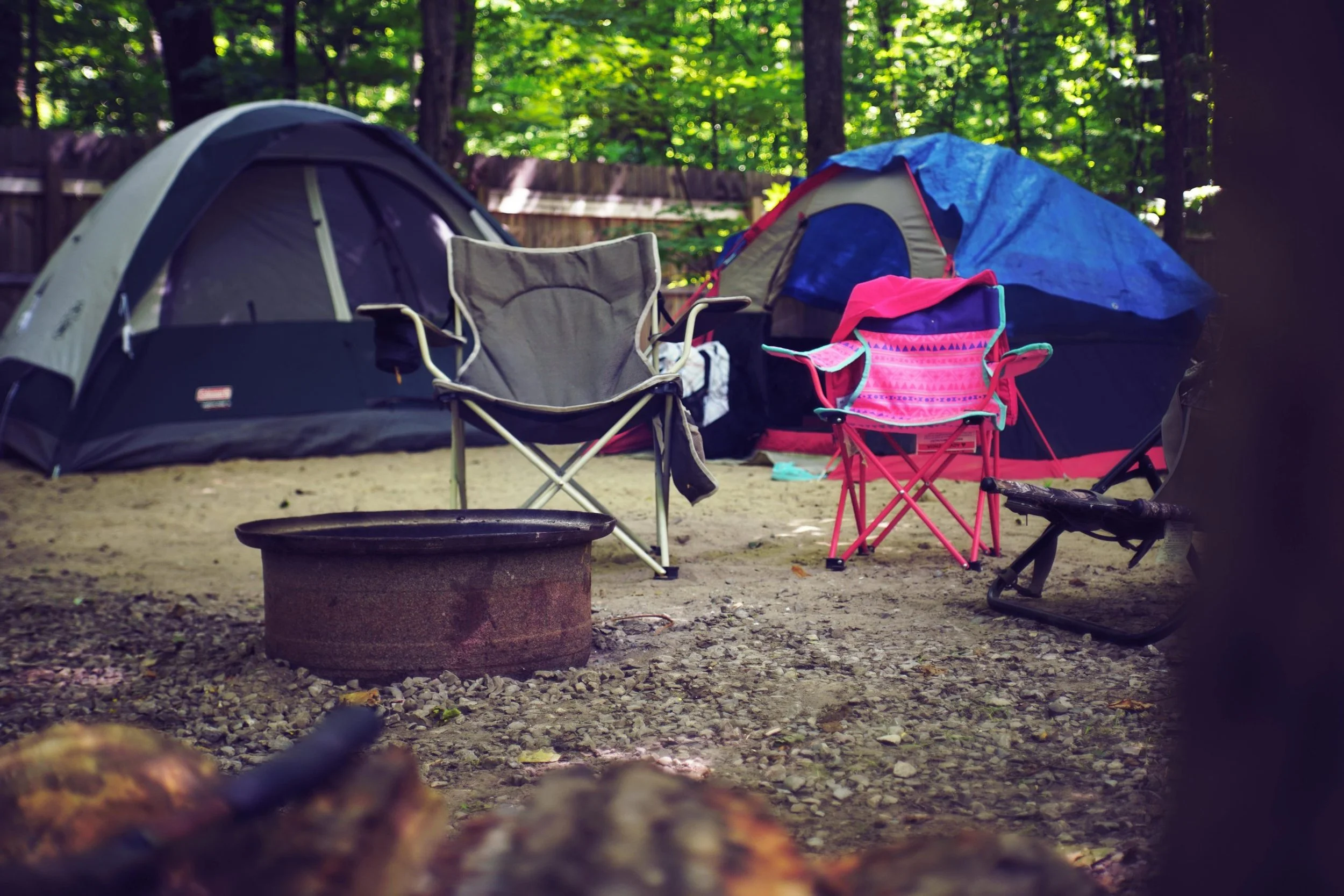 Camping scene in a wooded area with tents, folding chairs, and camping gear. The scene shows two tents, one large and one smaller, with a forest background. There is a foldable chair, a pink camping chair, and some miscellaneous camping items visible on the ground.