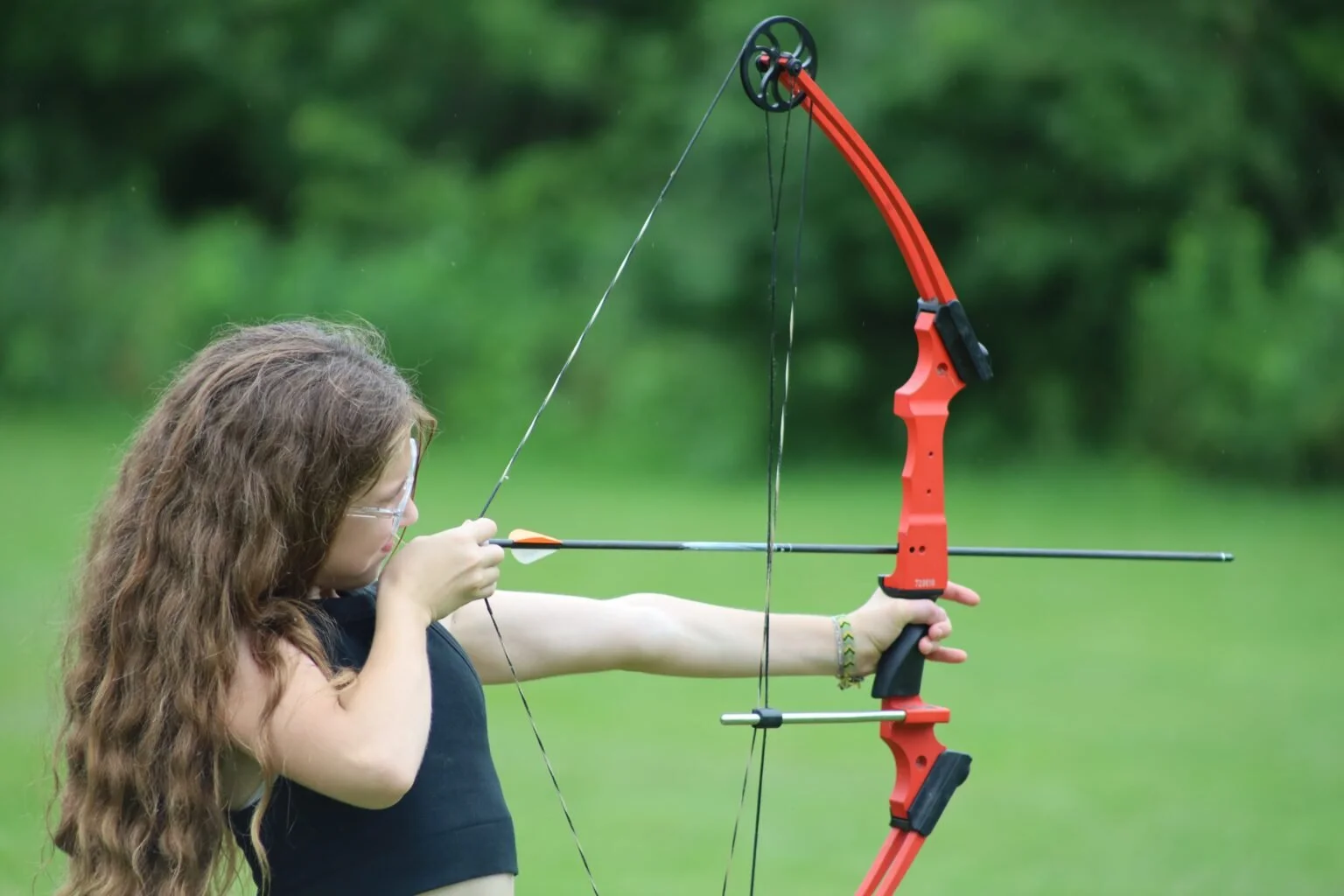 Girl with long curly hair aiming a red and black archery bow outdoors on a grassy field.