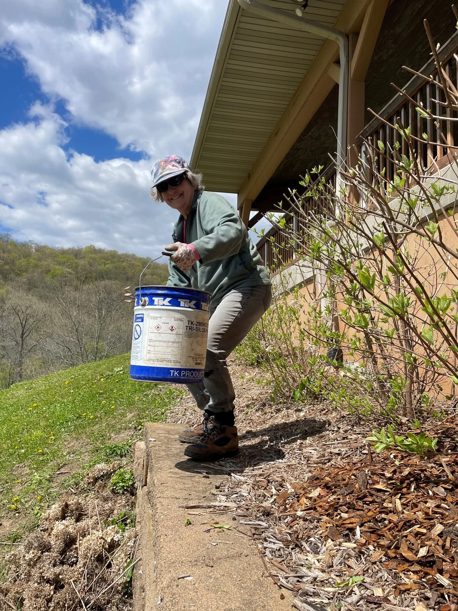 Woman with gray hair, sunglasses, colorful hat, gloves, and outdoor clothing holding a bucket of paint while standing outdoors near a house with a covered porch, on a sunny day with clouds in the sky.