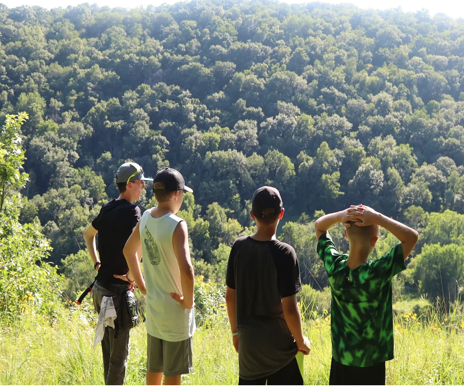 Four boys standing in a grassy field looking at a wooded hillside in the distance. One boy has his hands on his head, and the others are standing with their hands on their hips or relaxed.