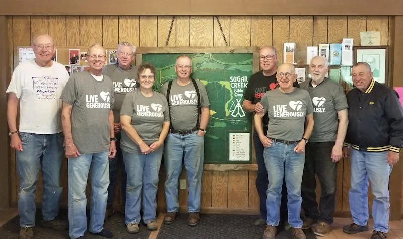 Group of nine people standing in front of a wooden wall with a Sugar Creek sign and display case, some wearing matching 'Live Generously' t-shirts.