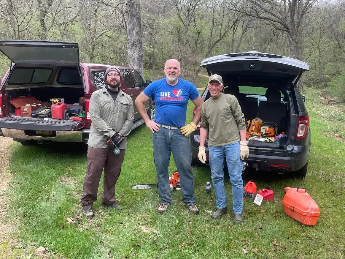 Three men standing outdoors in a grassy area with trees in the background. Two of them are wearing work gloves, and they are surrounded by open vehicle trunks filled with tools and equipment, suggesting they are doing outdoor work or repairs.