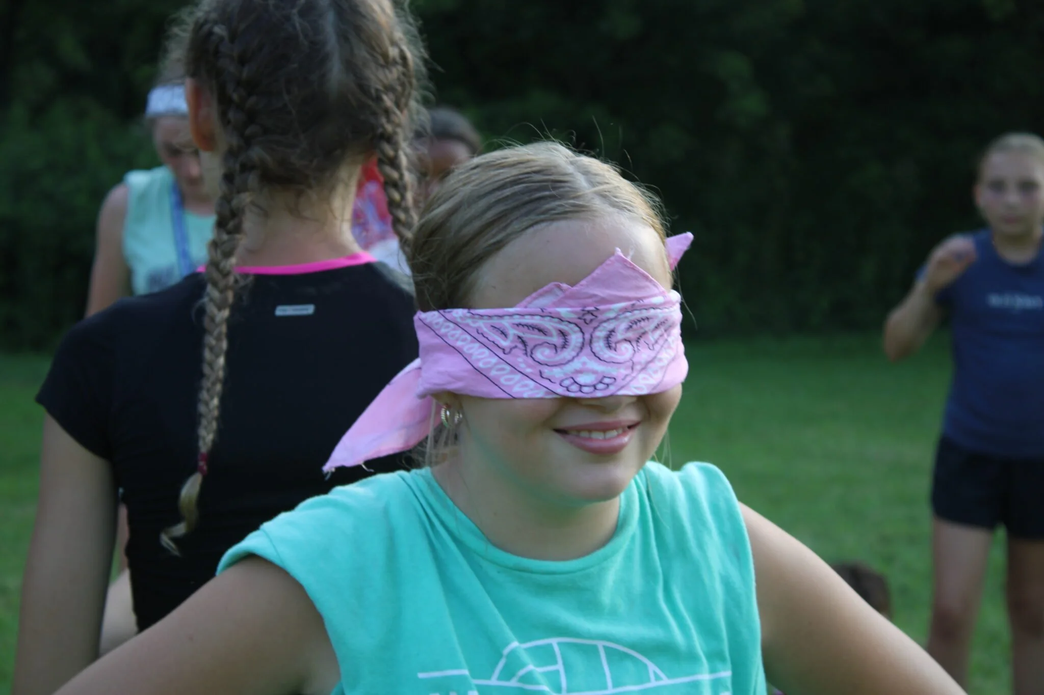 Group of children outdoors playing, one girl in the foreground with a pink bandana over her eyes and a light blue shirt, smiling, while others are in the background