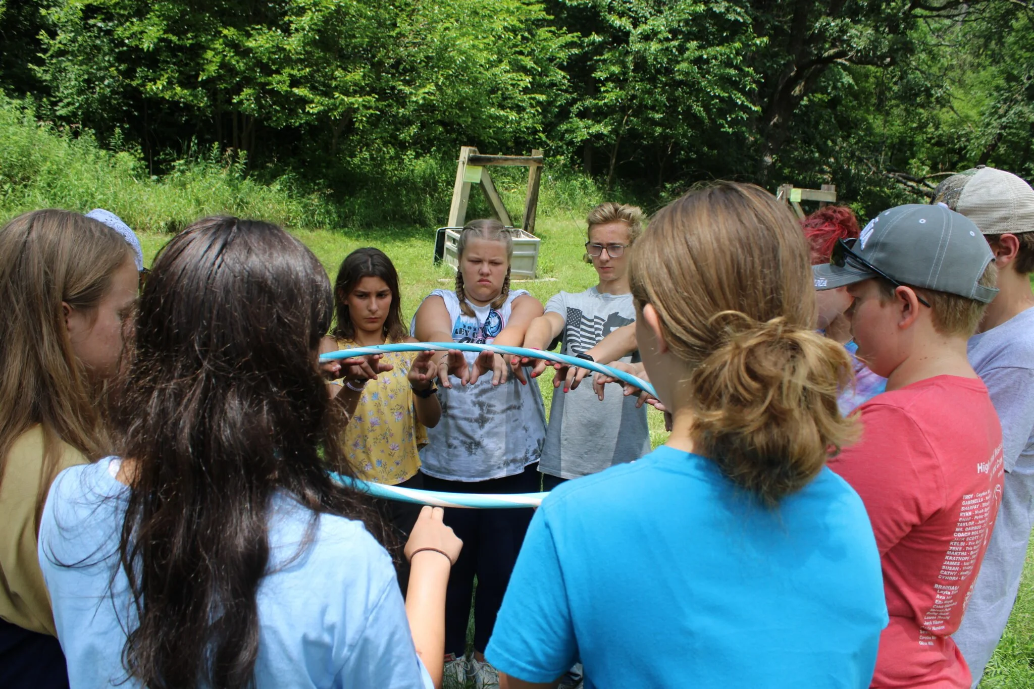 A group of children and teens standing outdoors in a circle, holding a hula hoop together. They appear to be participating in an outdoor activity or team-building exercise, with a forested background.