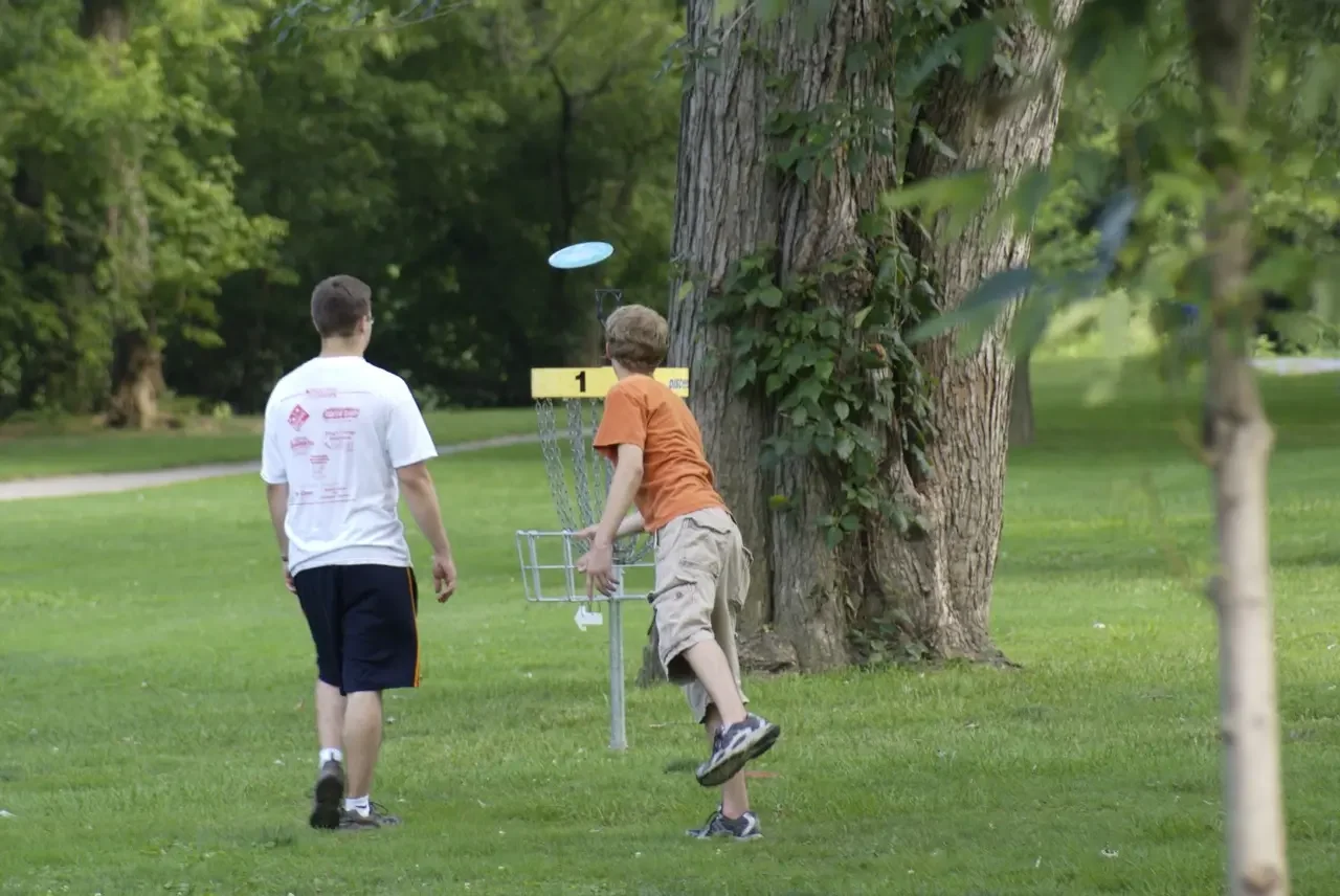 Two boys playing disc golf outdoors in a park, with one throwing a disc towards the disc golf basket.