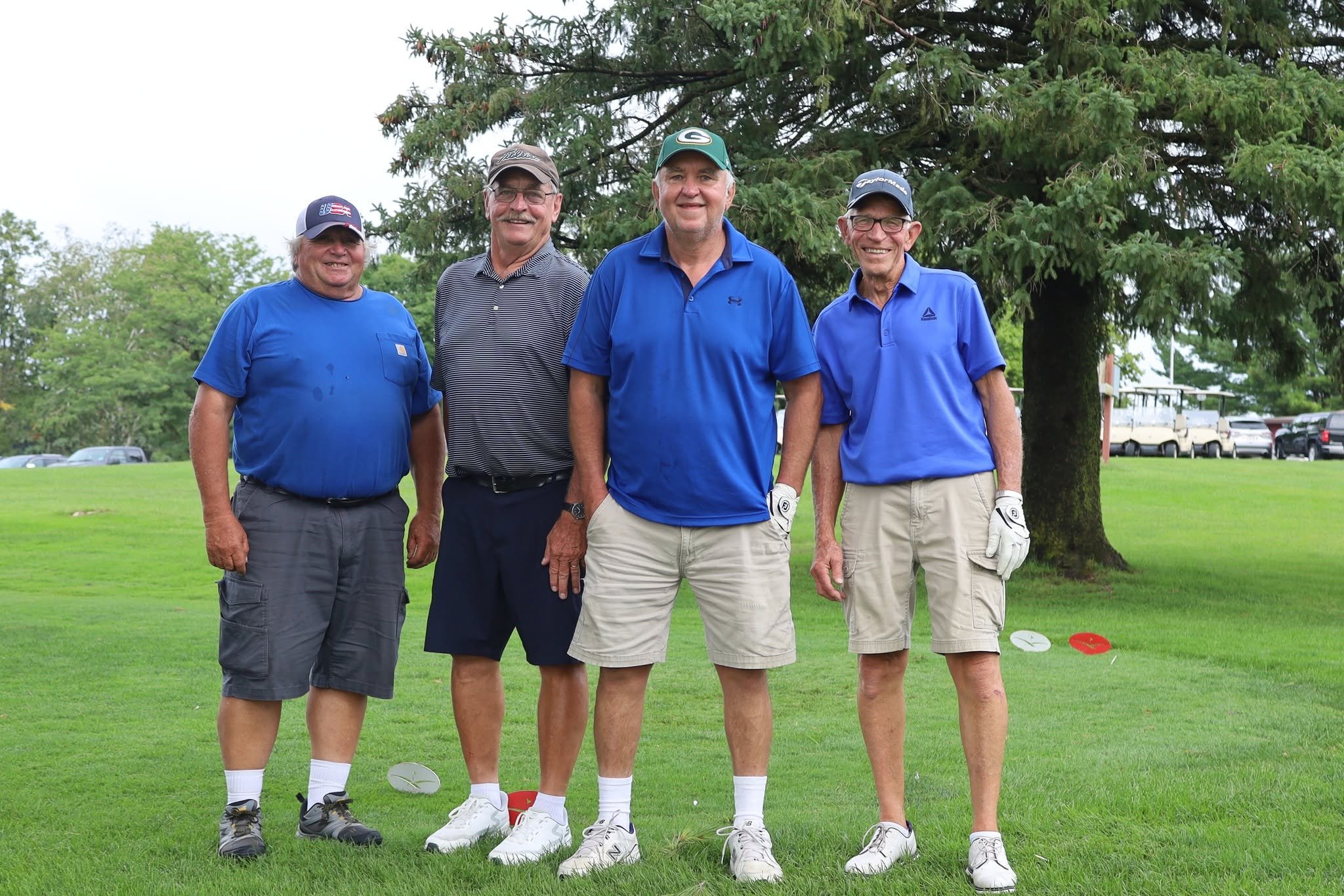 Four middle-aged men standing outdoors on a golf course. They are dressed in golf attire, including caps, golf gloves, and shorts. They are smiling and appear to be enjoying their time together.