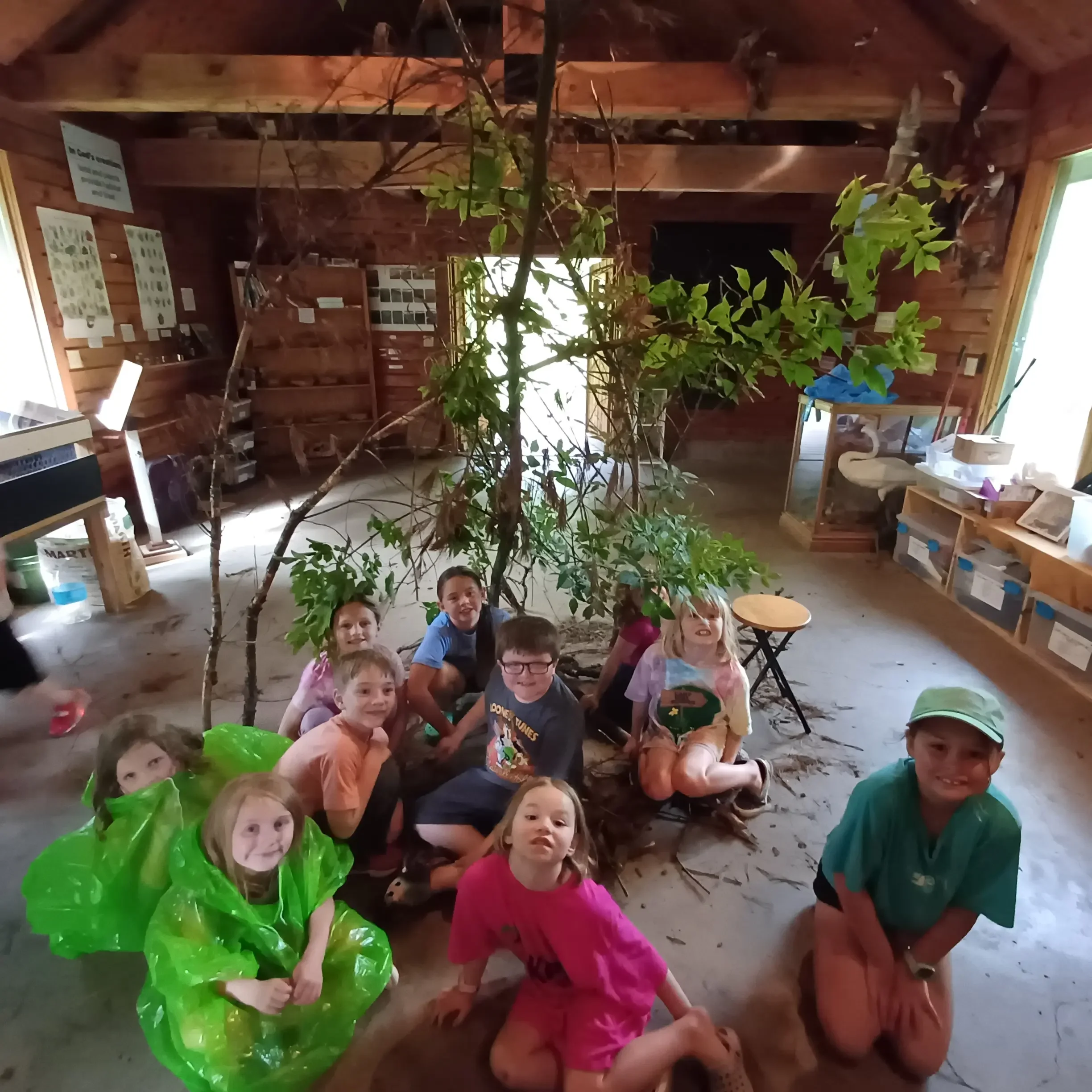 Group of children sitting on the floor inside a wooden building, smiling, with a large green leafy plant in the foreground.