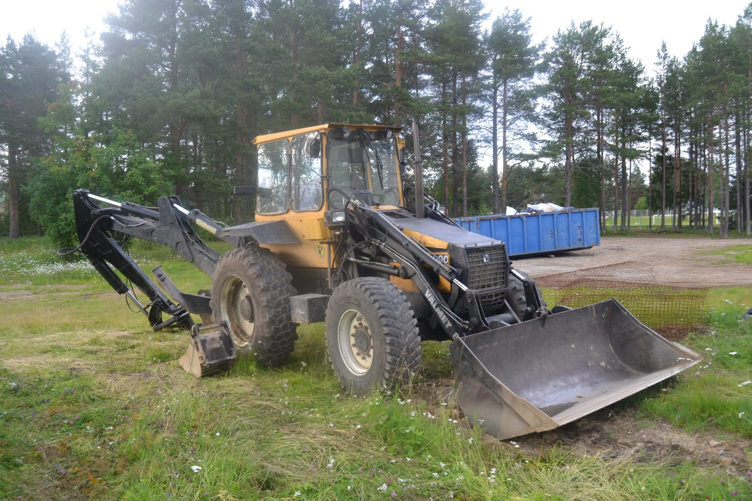 A yellow and black bulldozer parked on grassy land, with a large blue container behind it, in front of a dense wooded area.