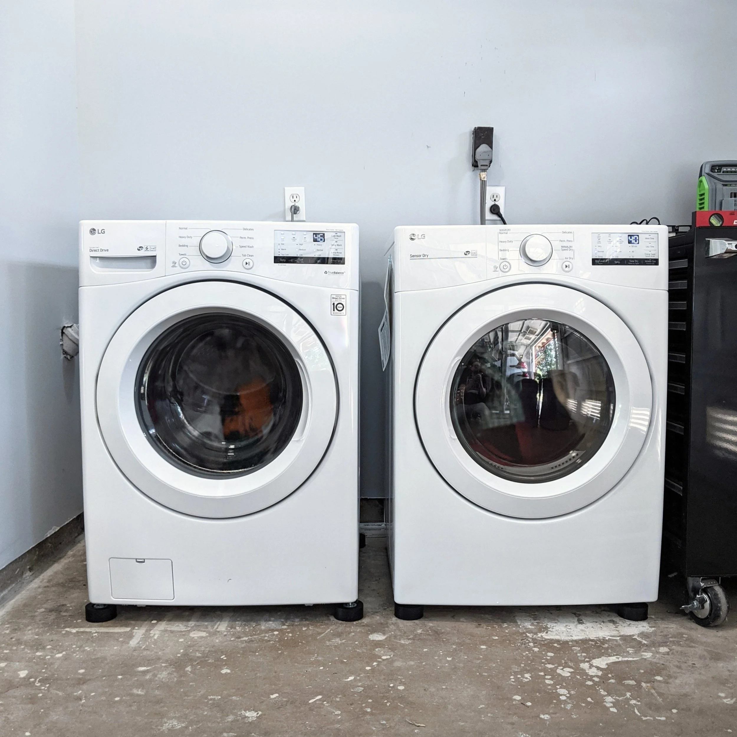 A laundry room with two white LG washing machines side by side, placed against a plain white wall. The machines have digital displays, and the floor is unfinished concrete with some debris.