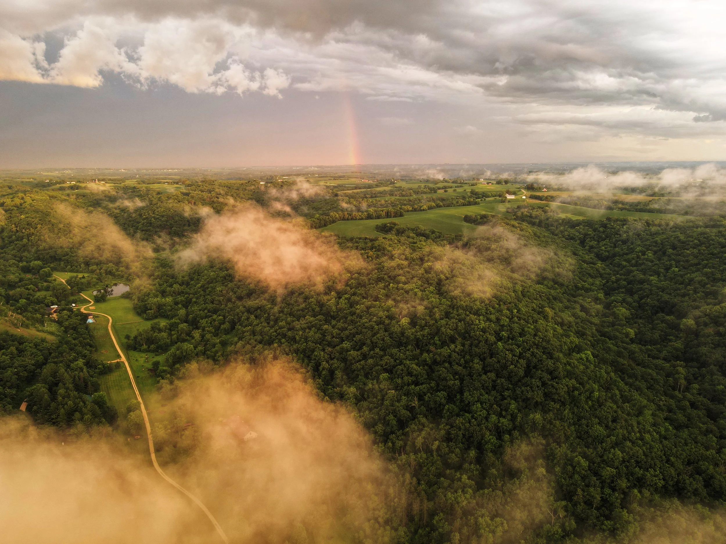 A landscape showing a green countryside with fields, trees, and a winding dirt road. There are patches of fog or low clouds and a rainbow faintly visible in the sky with dark clouds overhead.
