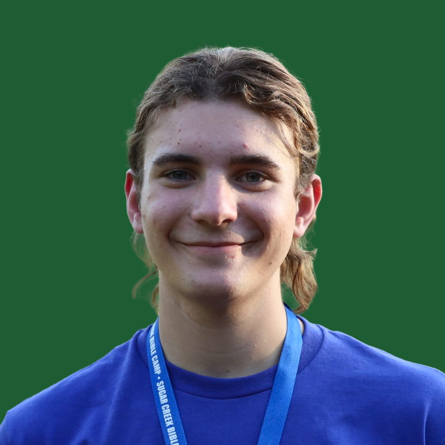 Young man with light brown hair, blue eyes, and freckles, wearing a blue shirt and a blue lanyard, smiling at the camera with a green background.