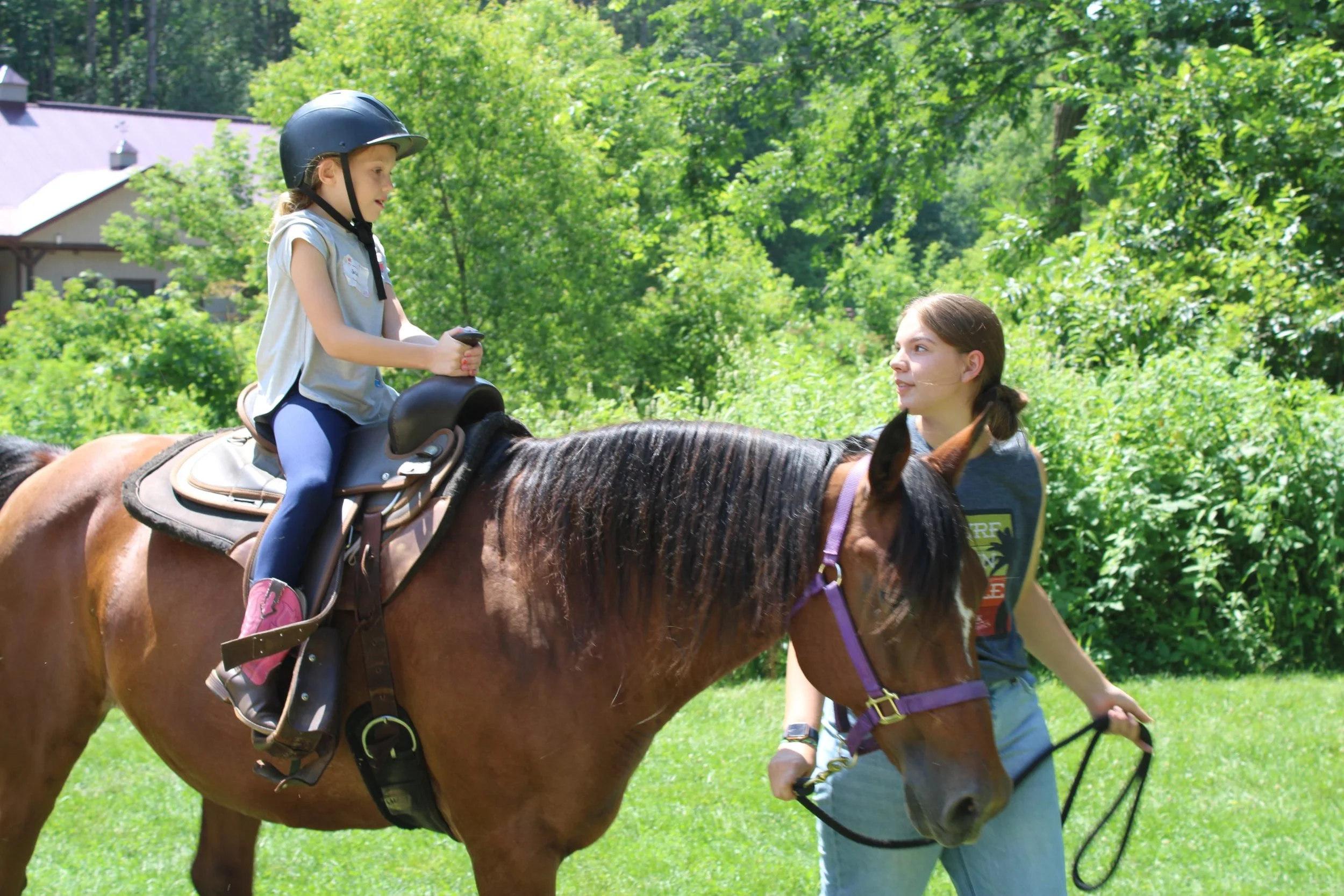 A young girl sits on a brown horse wearing a black riding helmet, with a woman walking alongside holding a lead rope. The girl appears focused as they are outdoors on green grass with lush trees in the background.