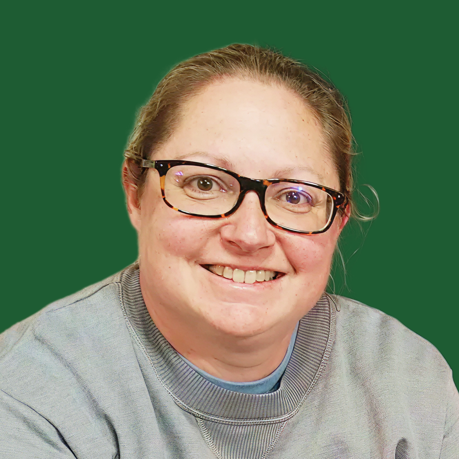 A smiling woman wearing glasses with a tortoiseshell pattern, a gray sweatshirt, and a blue shirt underneath. She has light brown hair and is seated against a plain green background.