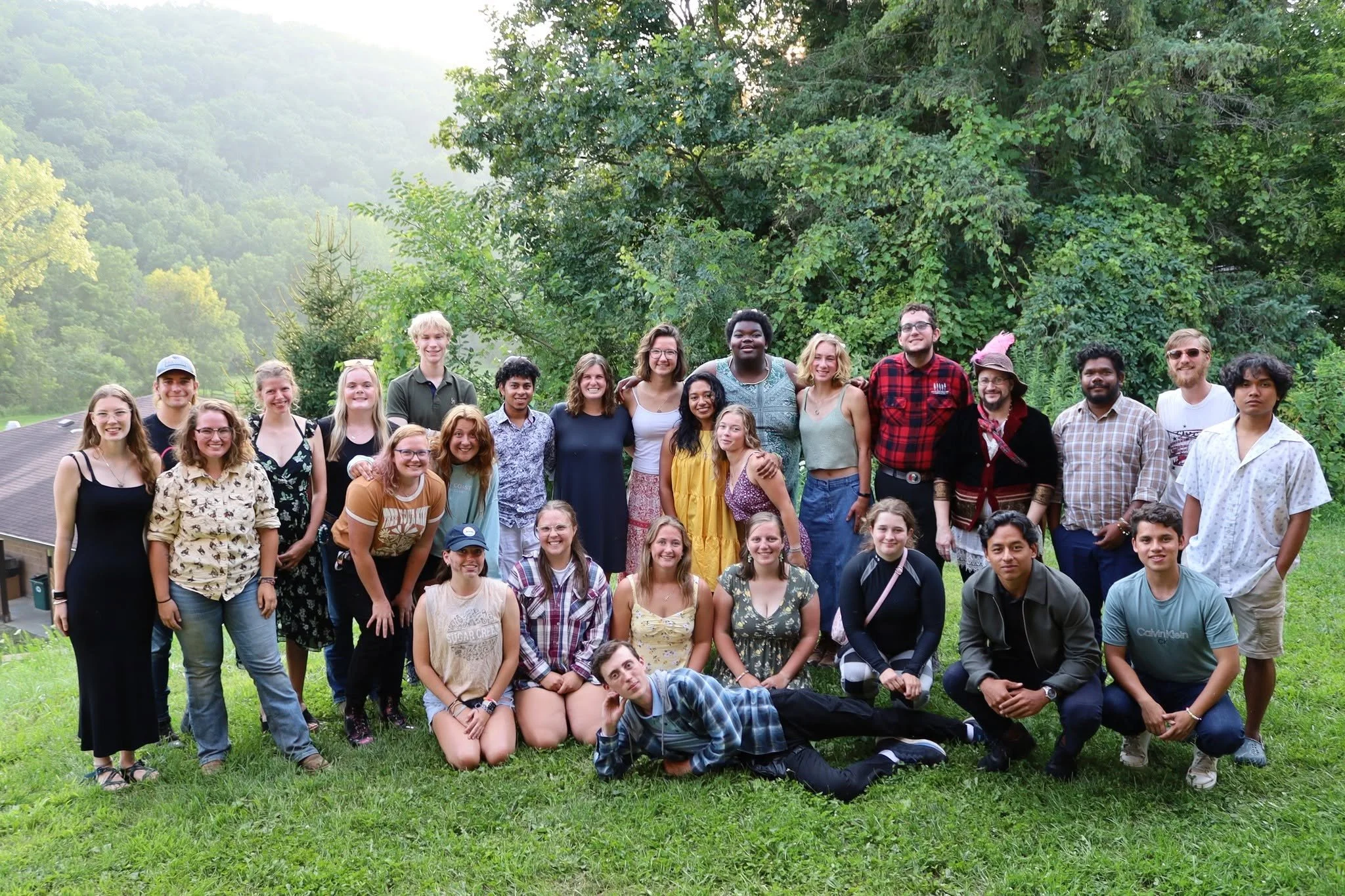 A large diverse group of young adults and teenagers posing outdoors on a grassy area with trees in the background, smiling at the camera.