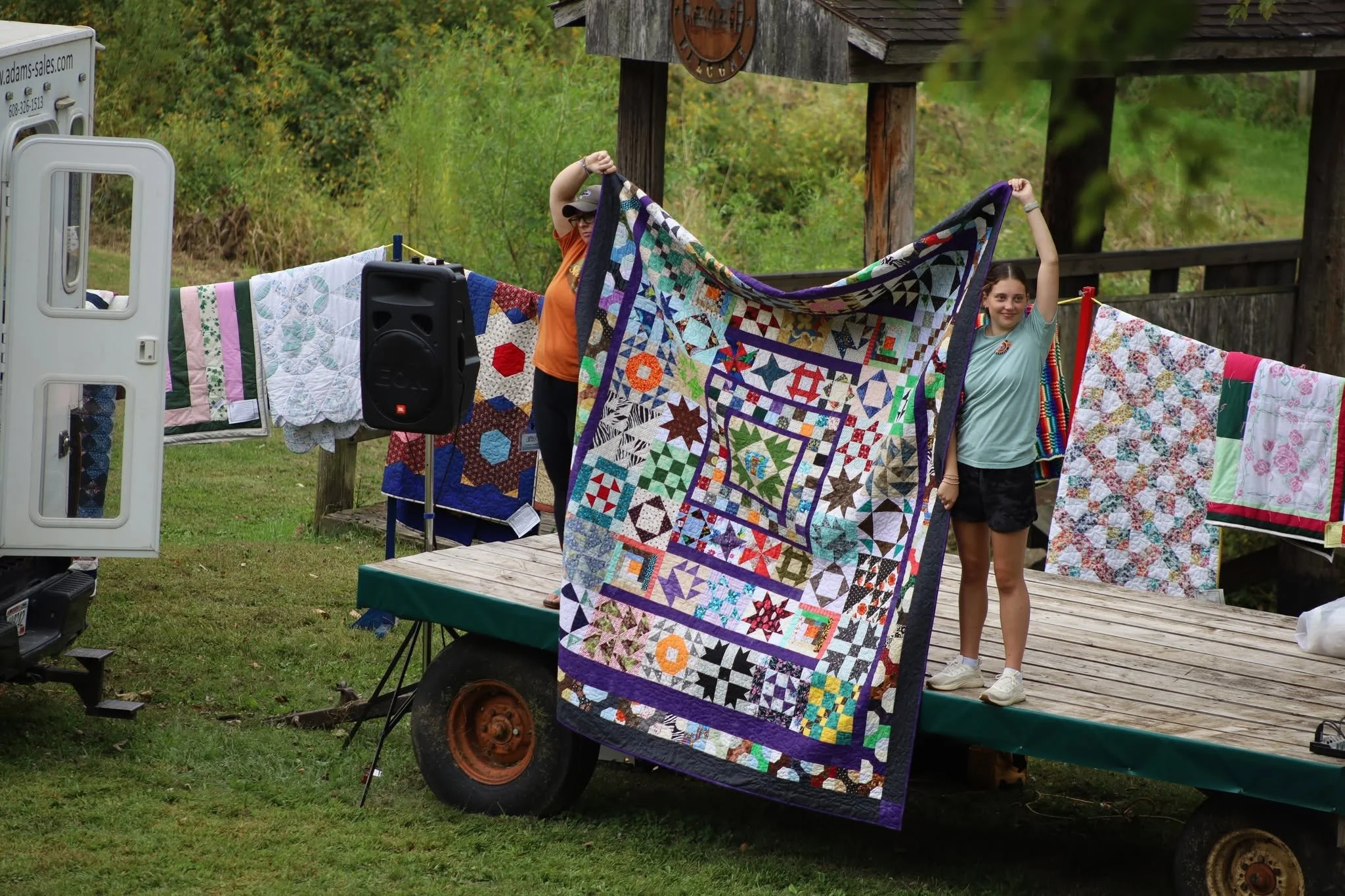 Two people hanging colorful and patterned quilts on a clothesline outdoors, with a wooden structure and lush green foliage in the background.