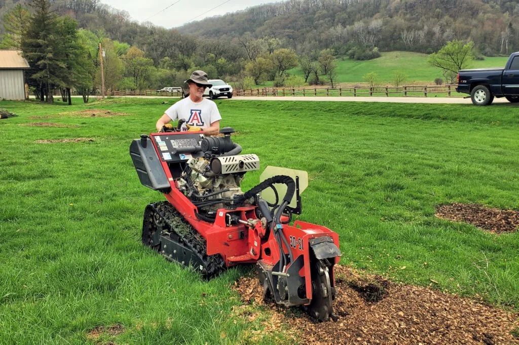 Person using a small tracked excavator or bulldozer on a grassy field, with trees, hills, parked cars, and a building in the background.