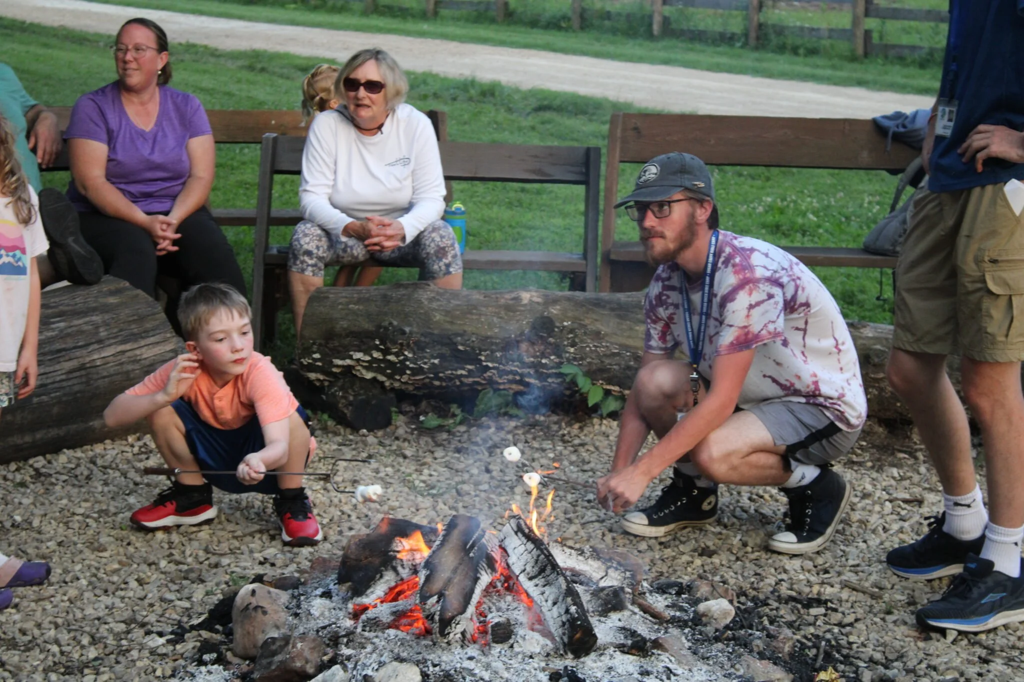 Group of people gathered outdoors around a campfire with logs and benches, some sitting and some standing, in a grassy area with a dirt road and fence in the background.