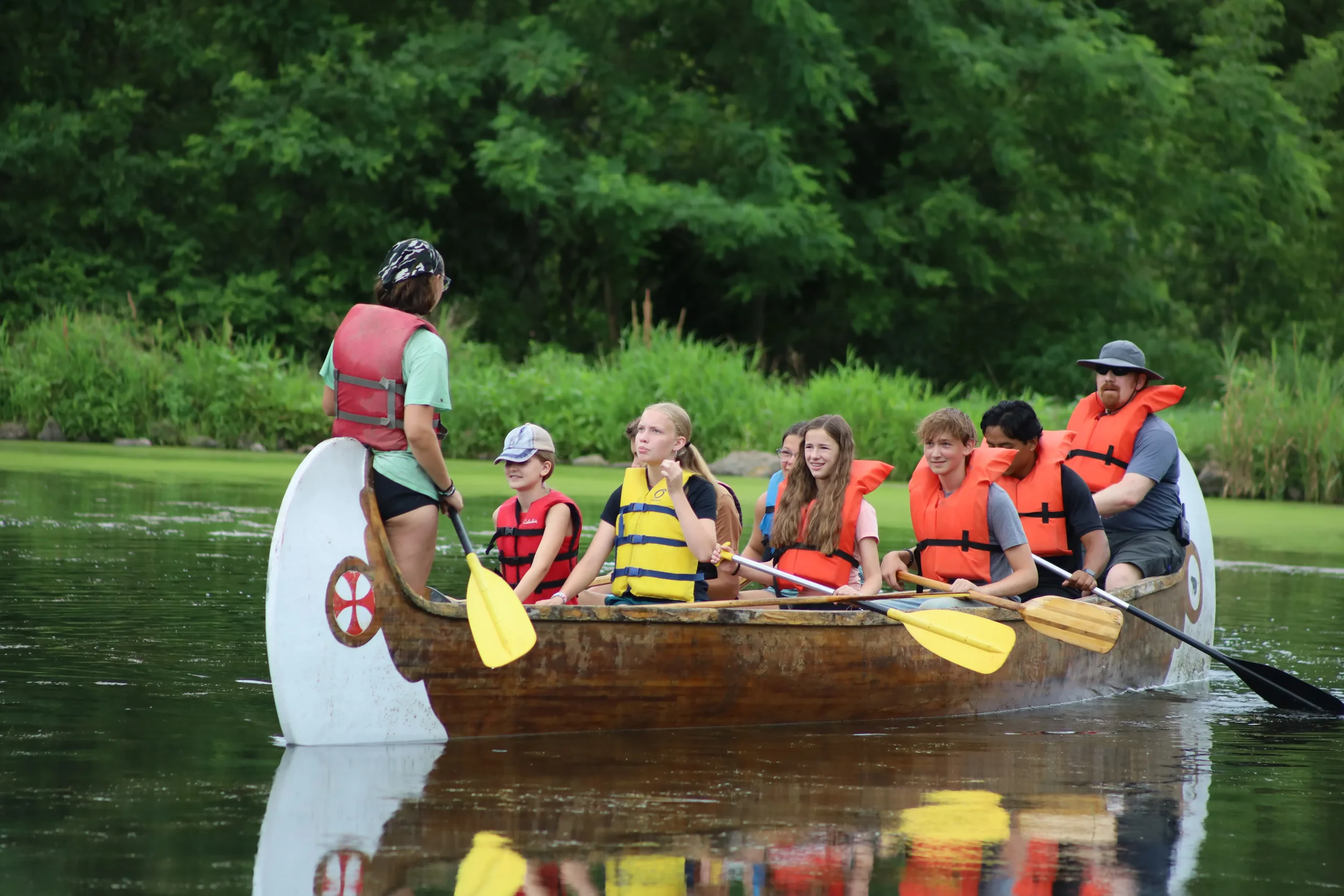 A group of children and an adult on a canoe on a calm river, surrounded by green trees, with everyone wearing life jackets and holding paddles, engaging in a water activity.