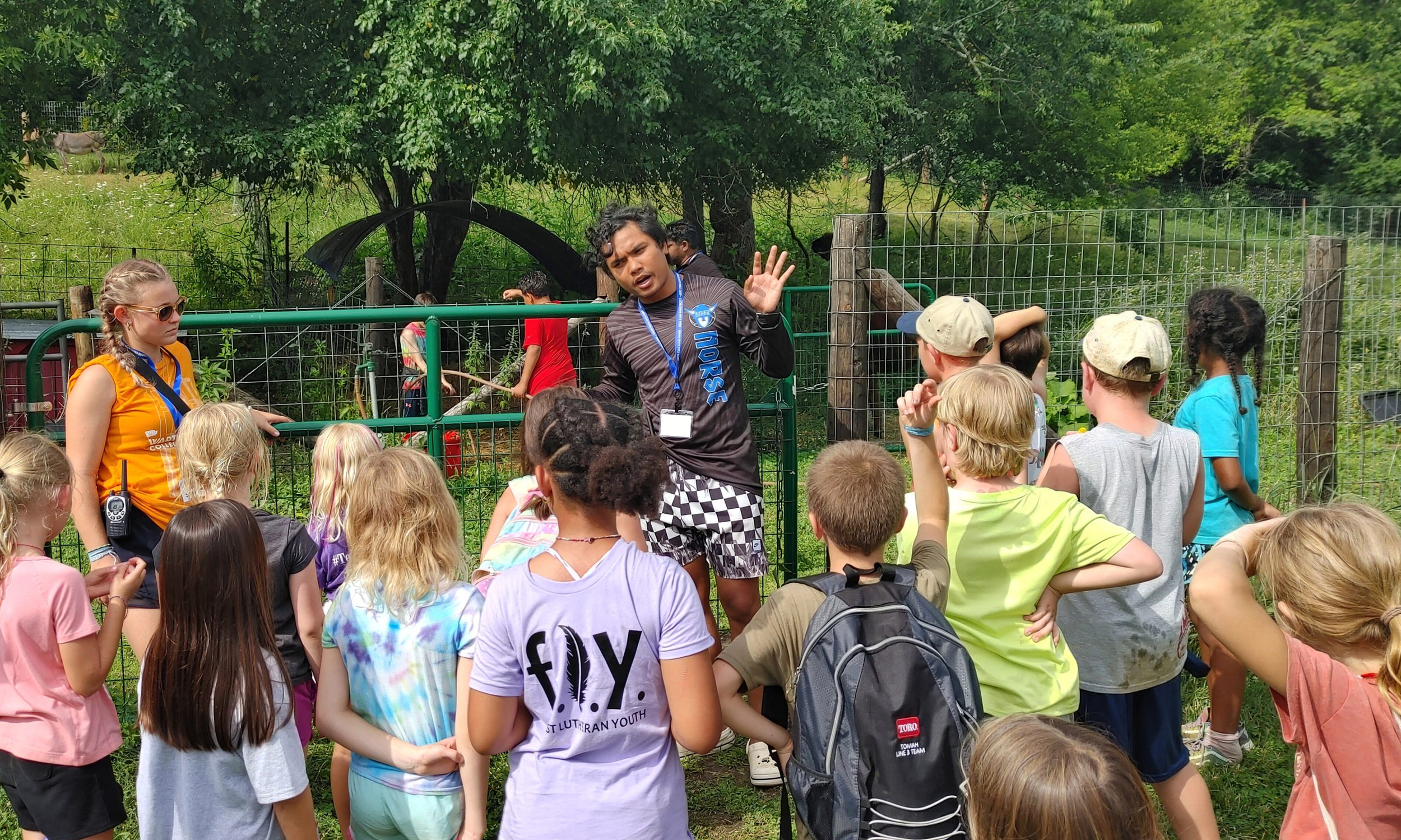 A group of children and a woman observing and listening to a man with a lanyard, in an outdoor setting with trees and a metal fence.