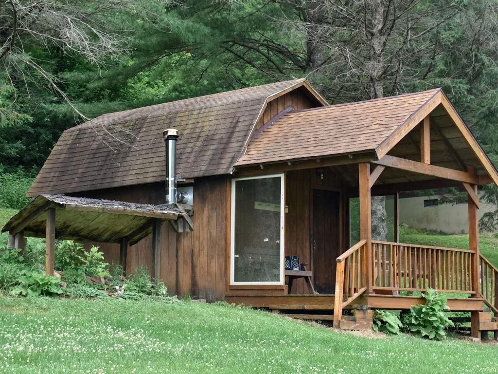 A small wooden cabin with a porch, surrounded by green grass and trees.
