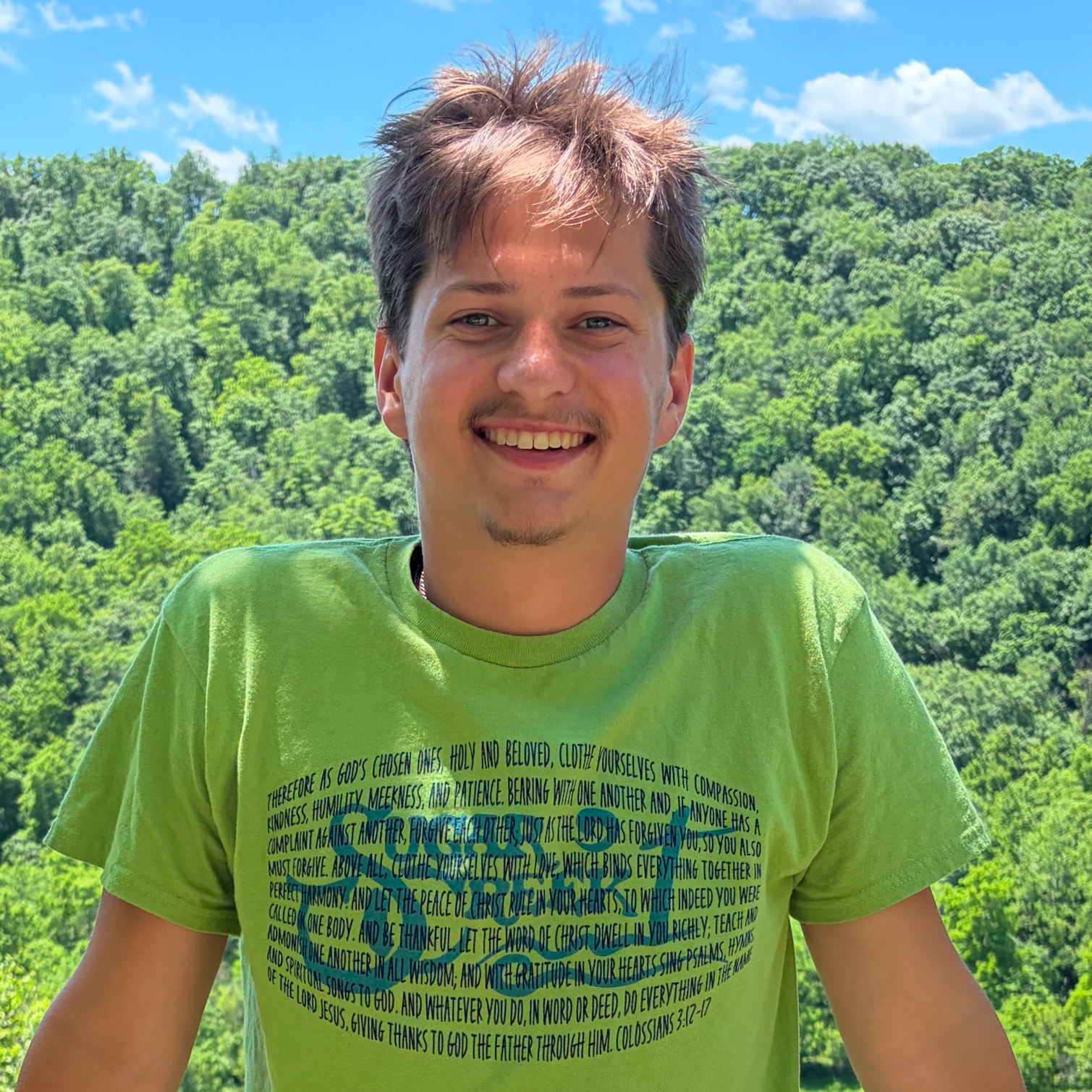 A young man with brown hair and a goatee smiling outdoors against a background of green trees, blue sky, and white clouds, wearing a lime green T-shirt with blue text.