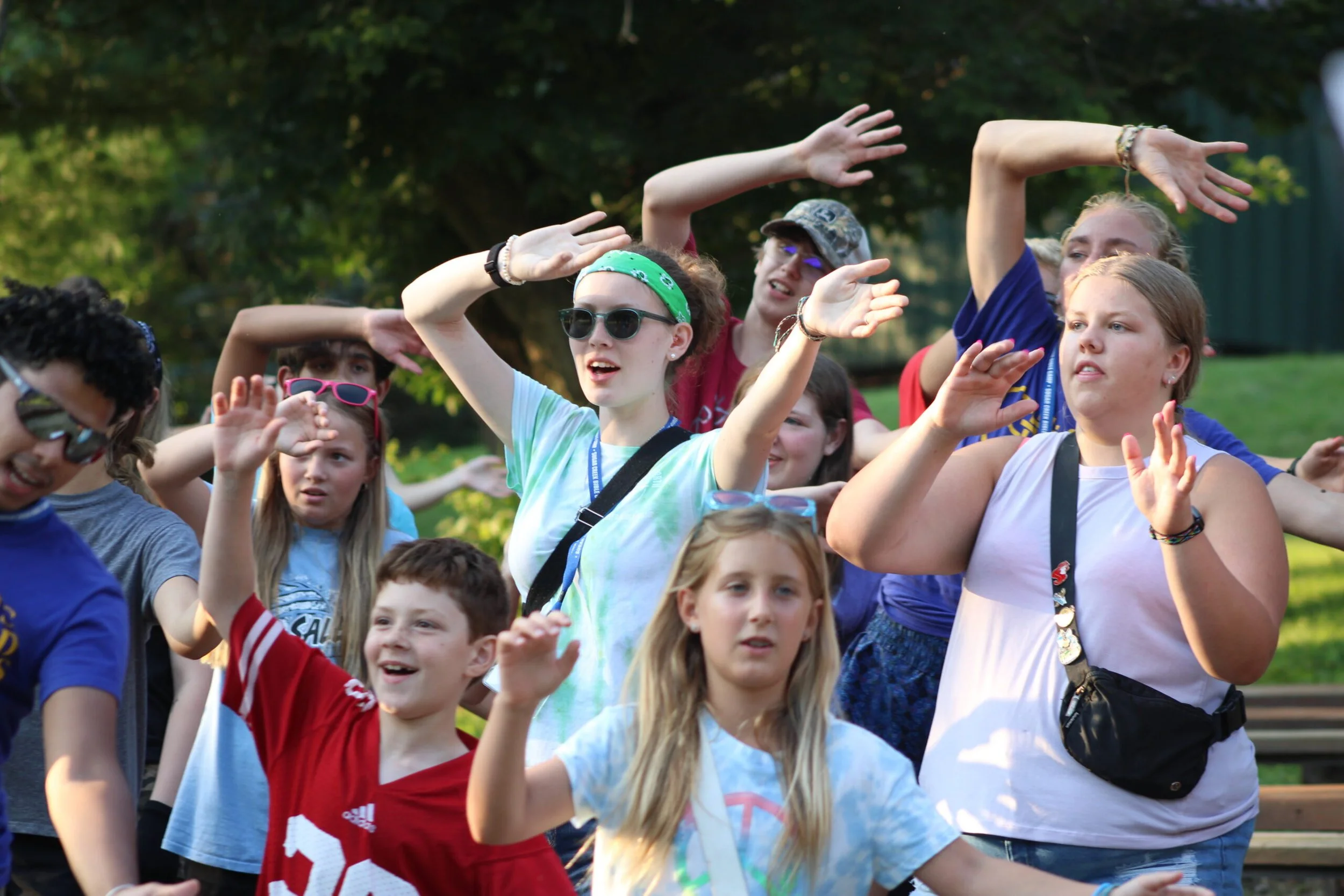 A group of children outdoors, wearing casual clothes and sunglasses, raising their hands in the air.