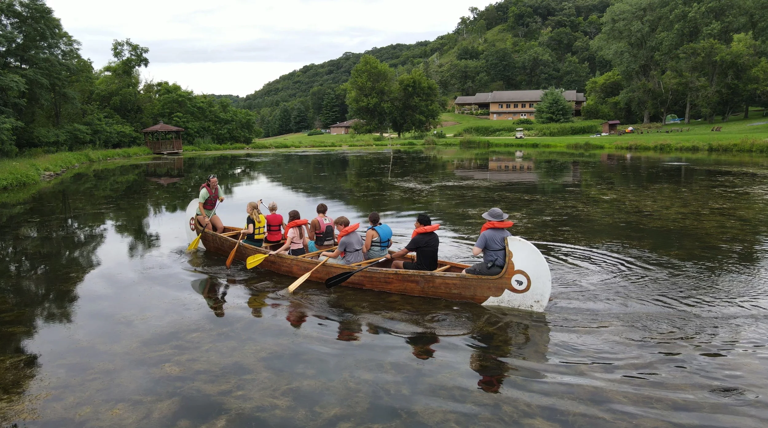 People in a dragon-shaped wooden boat paddling on a calm river surrounded by lush green trees and grass, with houses and a gazebo on the hillside in the background.