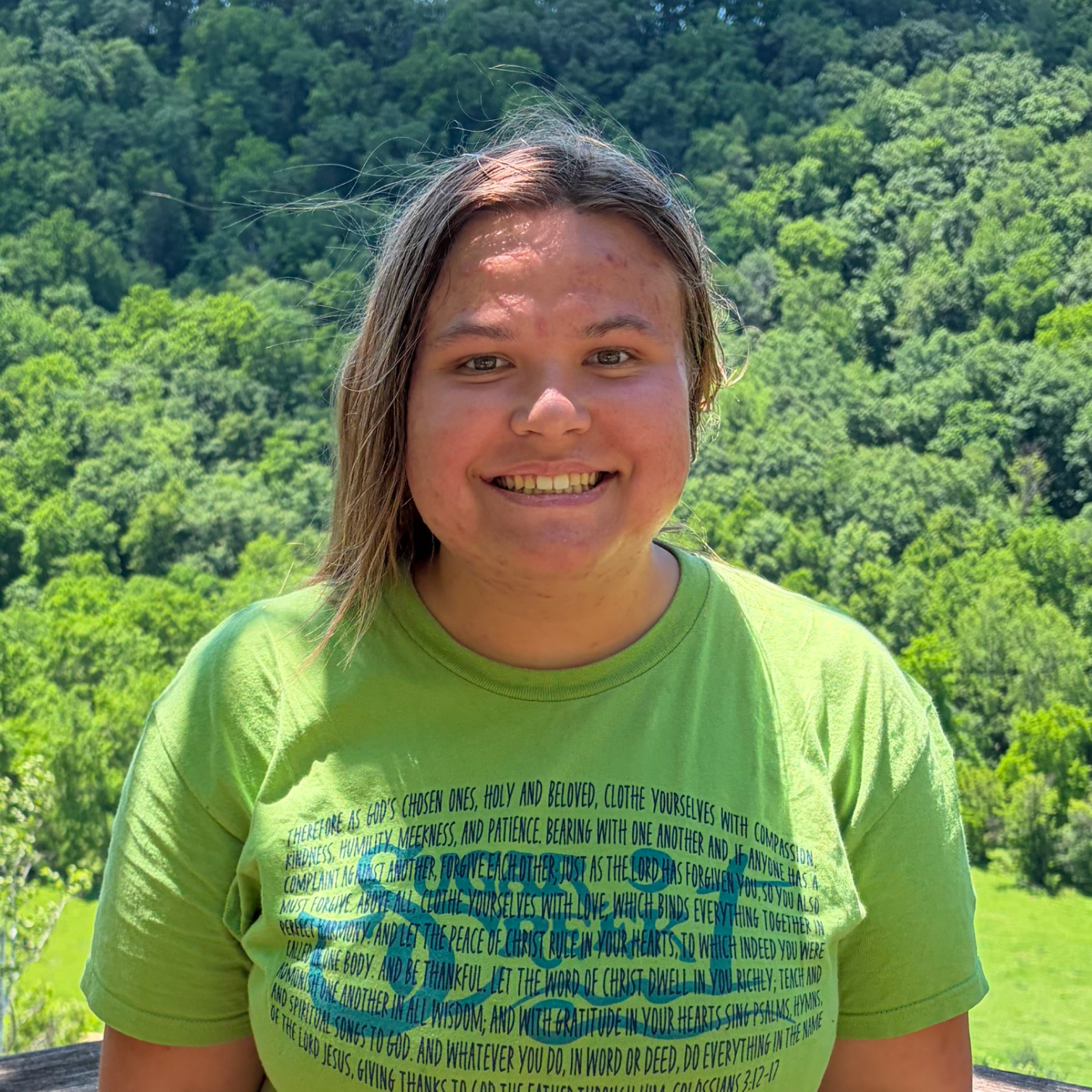 A person with long hair and wearing a bright green T-shirt, smiling at the camera outdoors with a lush, green forested hillside in the background.